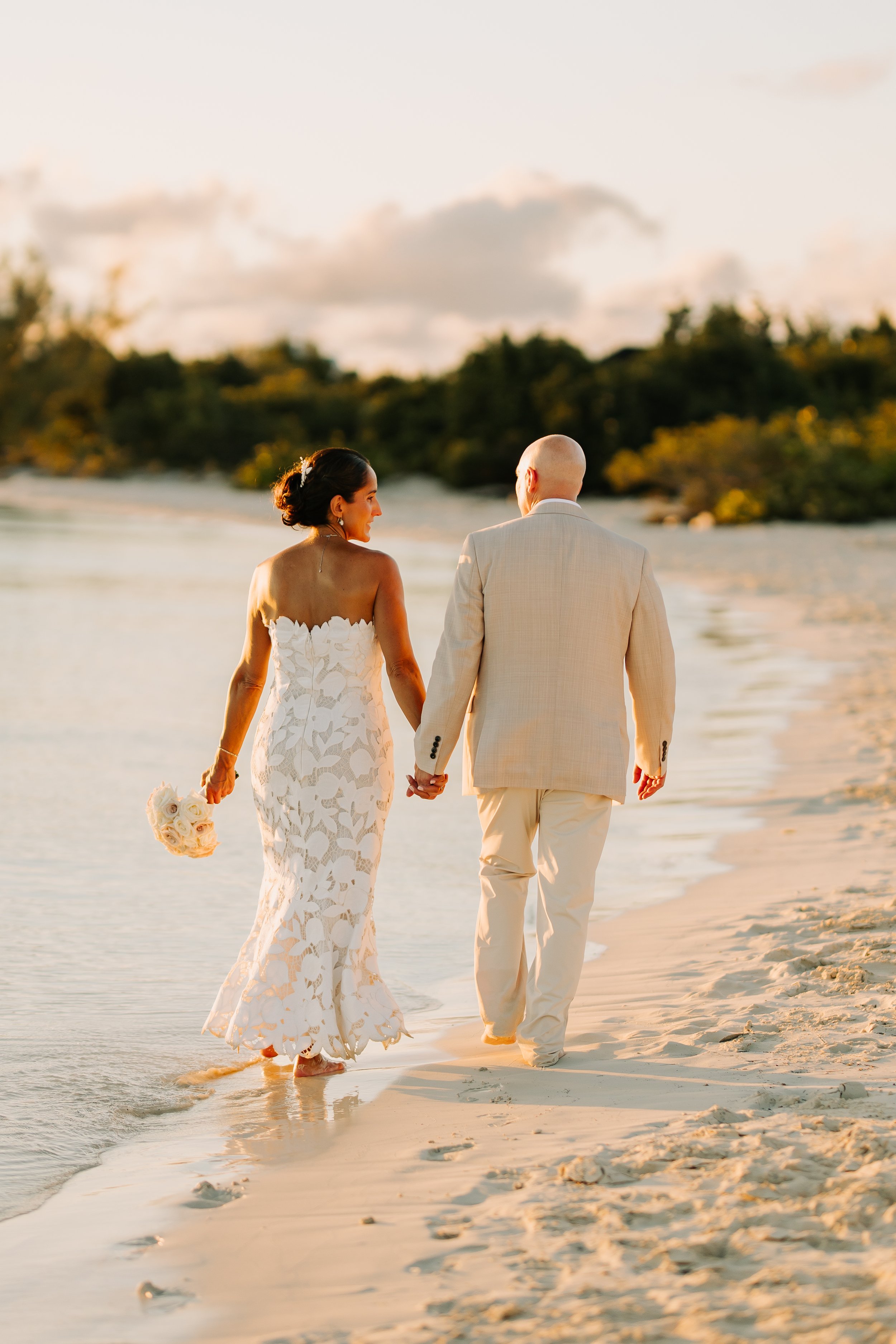 A bride and groom walking hand in hand along the beach during sunset, with the bride holding a bouquet of white roses and wearing a white lace wedding dress, and the groom dressed in a light-colored suit.