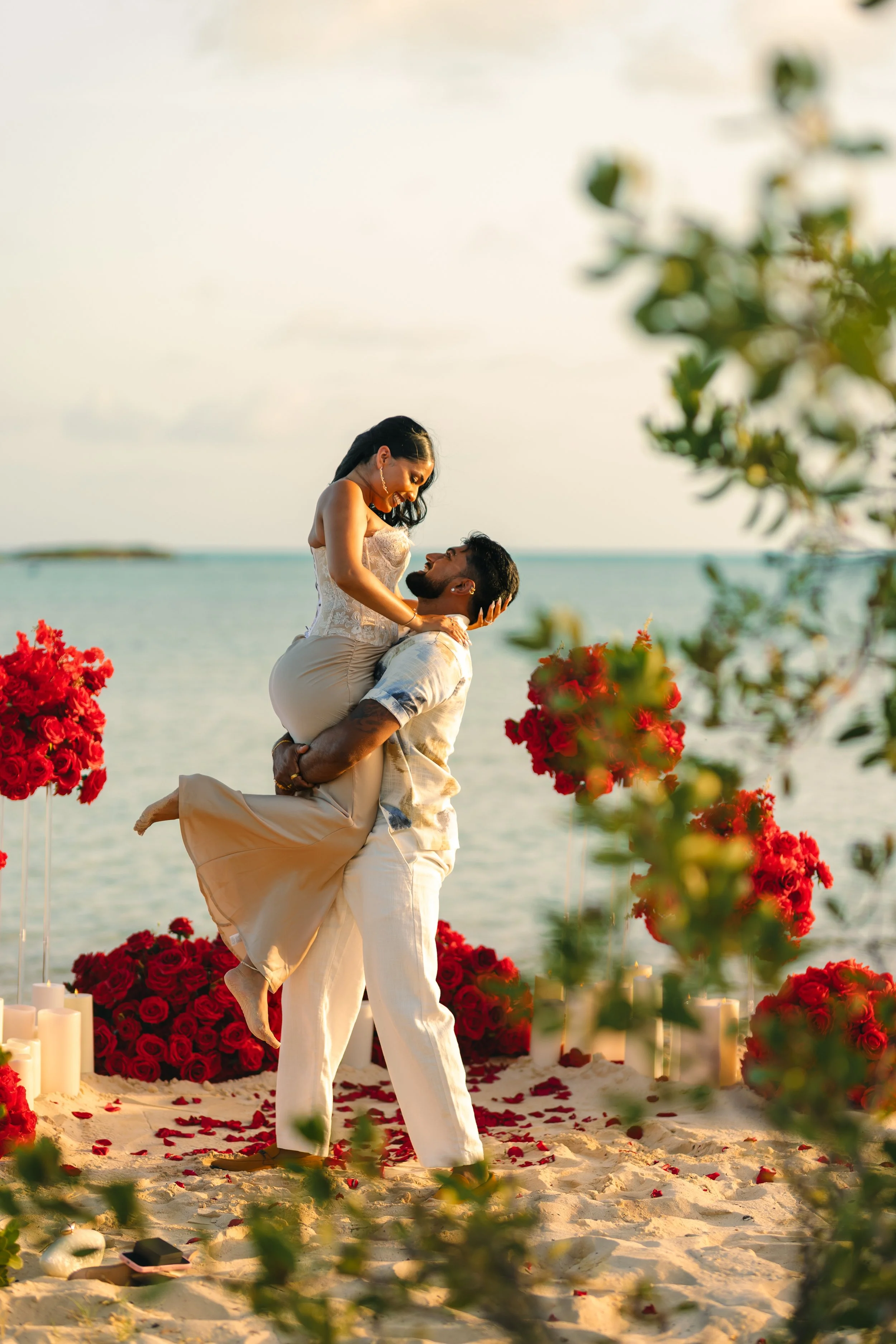 A couple is embracing on a beach during sunset, surrounded by red roses and candles, with the ocean in the background.