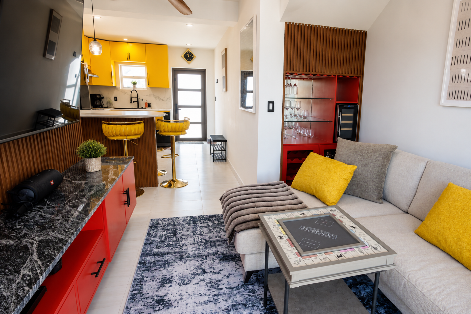 Living room with beige sofa with yellow and gray pillows, coffee table, gray and white rug, and a gray wall-mounted TV. Adjacent is a kitchen area with yellow cabinets, black countertop, and two yellow bar stools. A red wine and glass display cabinet