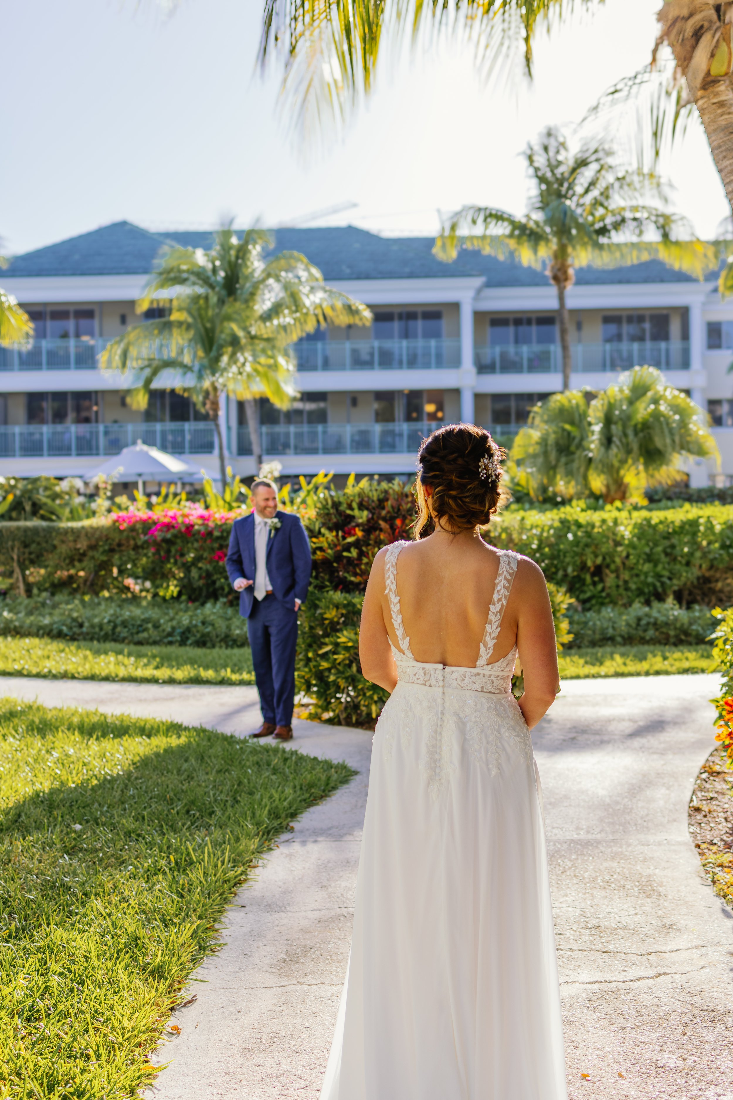 A bride in a wedding dress facing a groom in a suit outdoors in a garden with tropical plants and a hotel building in the background.