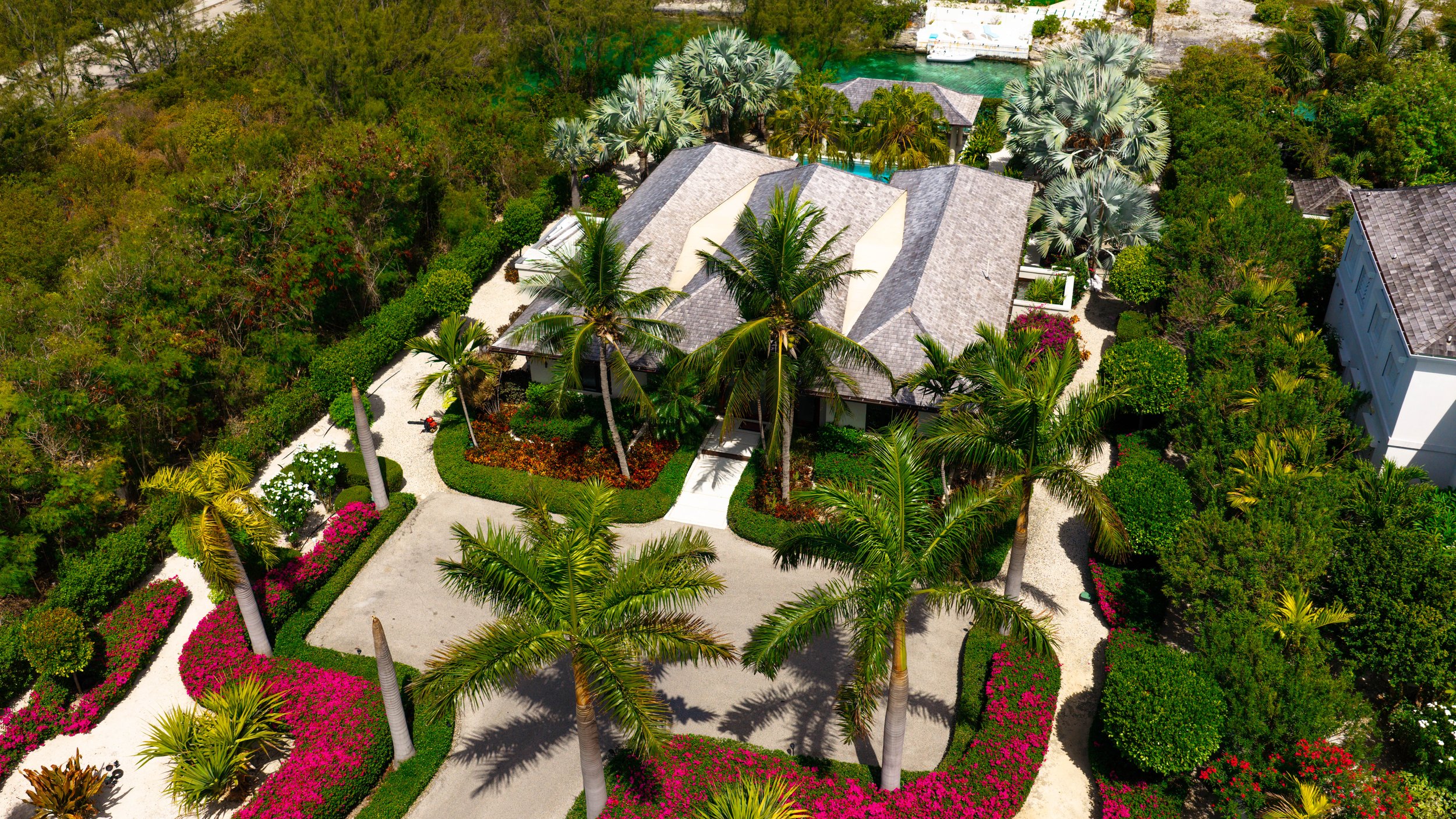 Aerial view of a house surrounded by lush greenery, palm trees, colorful flower gardens, and a winding pathway leading to the entrance, with a view of a waterway with a boat in the background.