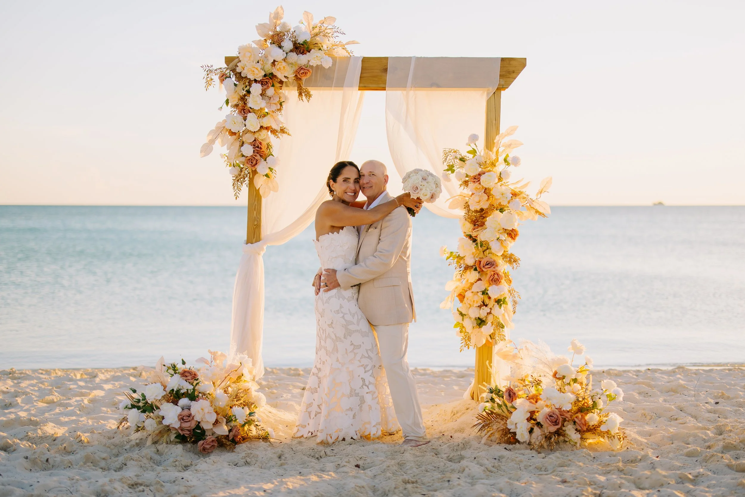 A newlywed couple stands on a sandy beach at sunset beneath a floral wedding arch decorated with cream, blush, and mauve flowers. The bride wears a white lace strapless gown and holds a bouquet of pale roses, while the groom wears a light beige suit. The calm ocean is visible in the background.