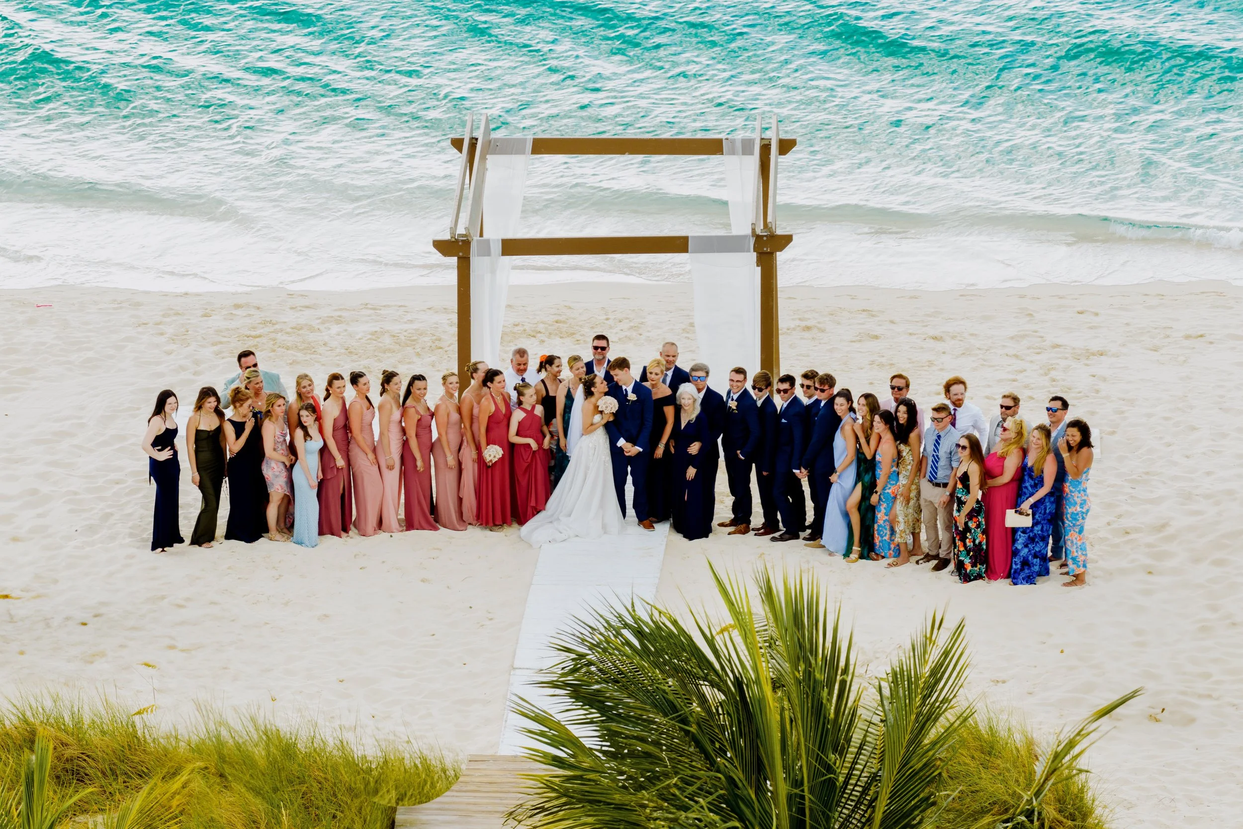 A large group of people, dressed in formal attire, gathered on a beach for a wedding photo. They are standing on a wooden pathway leading to a beach with the ocean in the background. The bride and groom are in the center with the bride holding a bouq