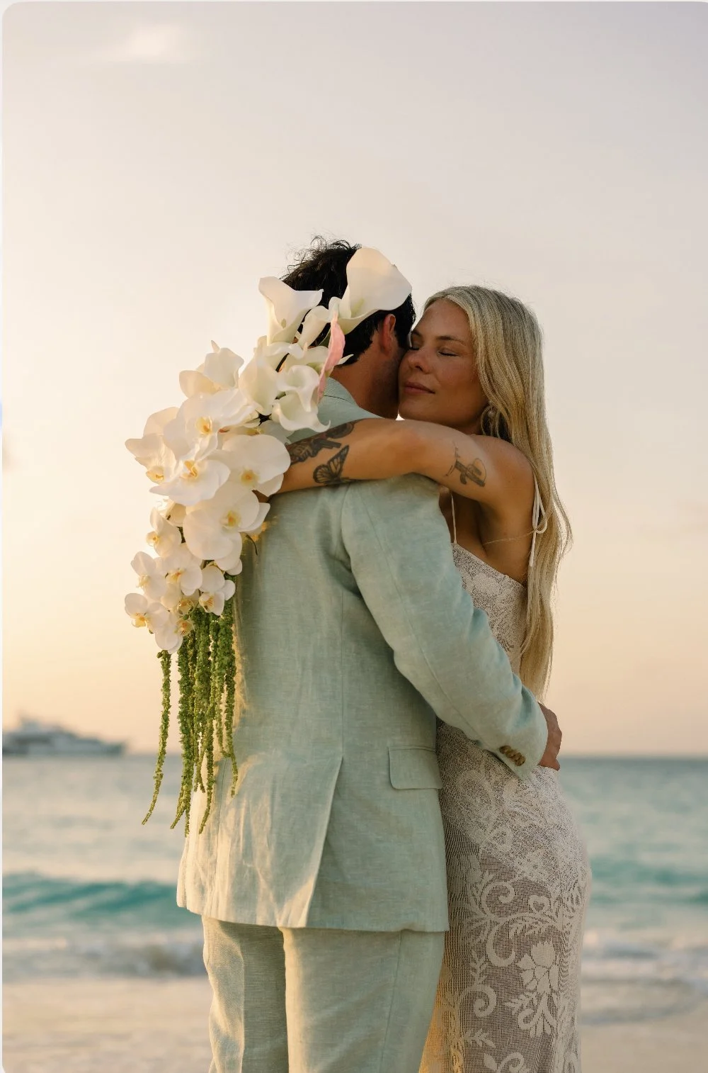 A couple embracing on the beach at sunset, with a bouquet of white orchids over the groom’s shoulder