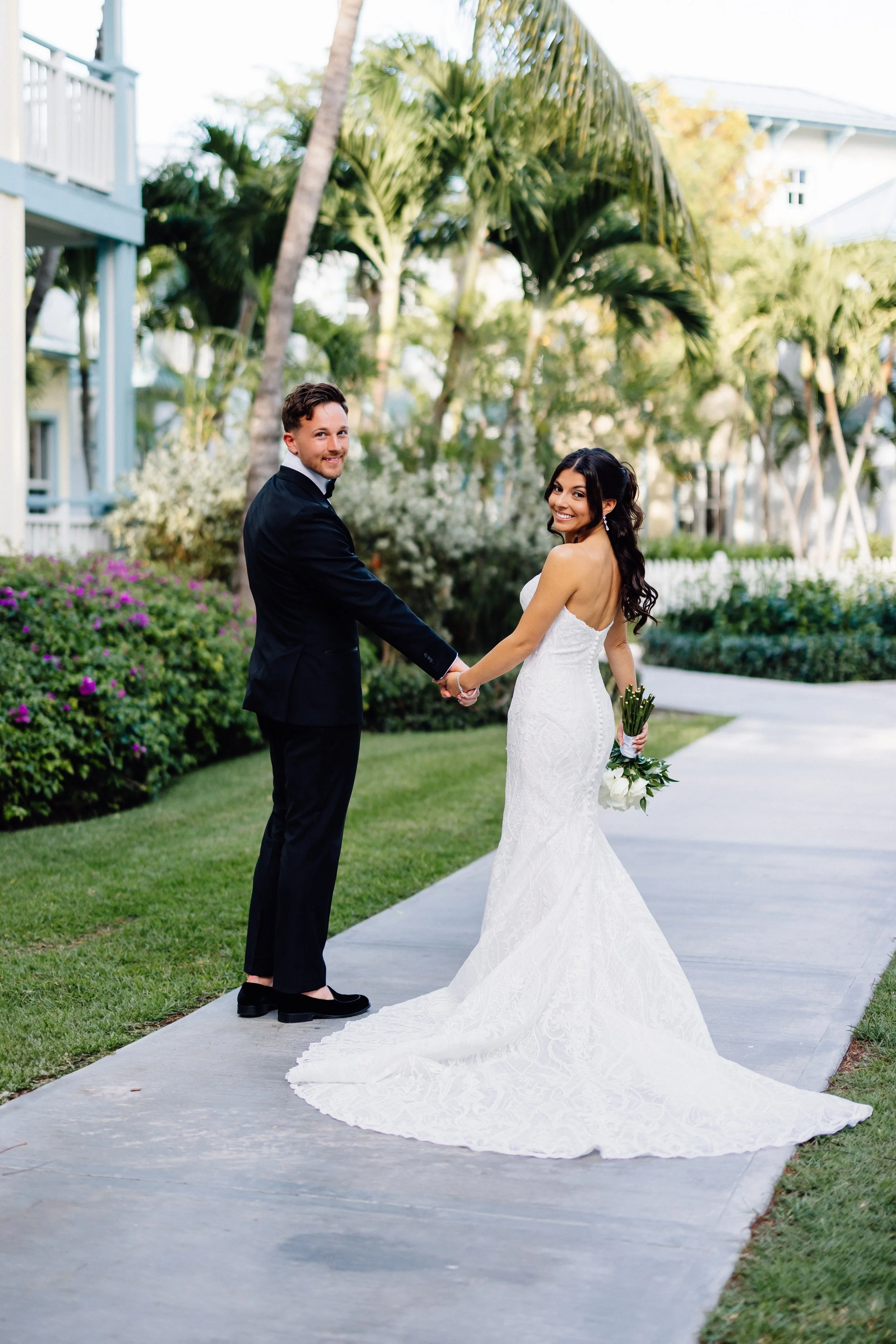 A newlywed couple holding hands and smiling outdoors near palm trees and a white building, with the bride in a strapless white wedding gown holding a bouquet of white flowers.