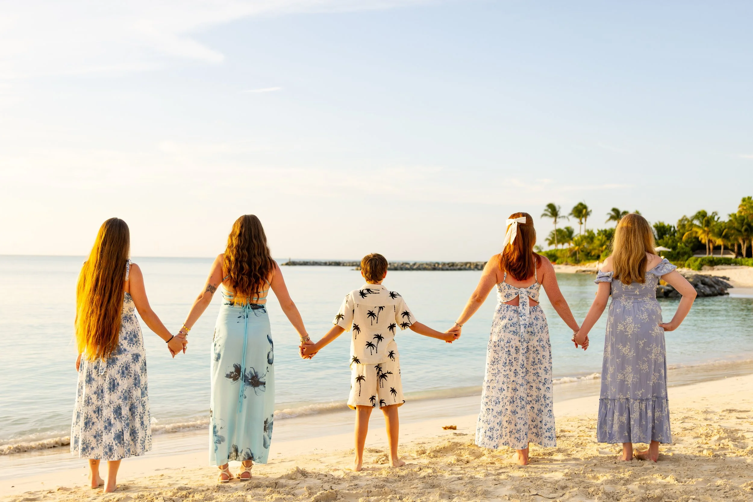 A group of five women and one young boy holding hands and standing on a sandy beach near the water, facing toward the ocean under a clear sky with palm trees in the background.