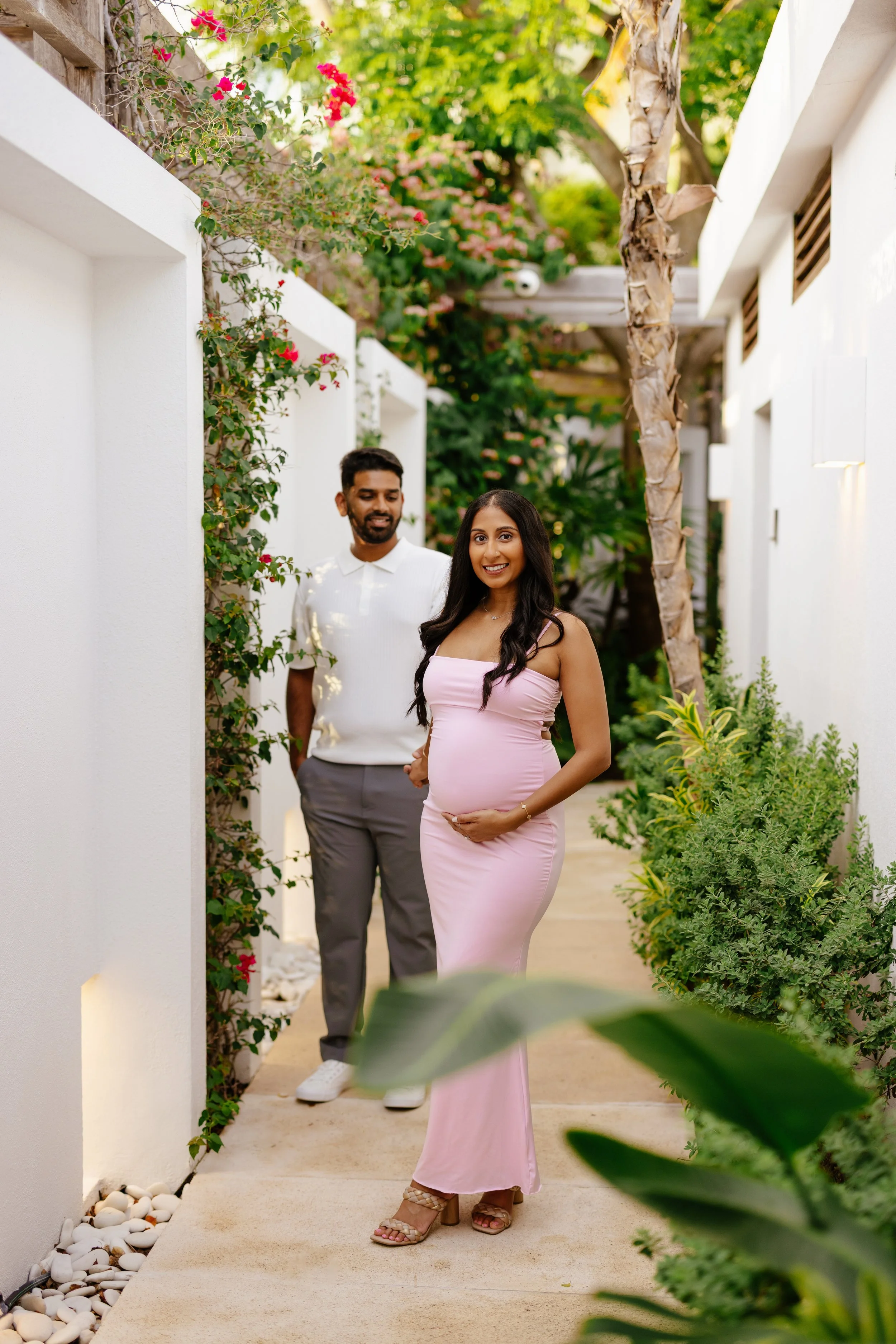 A pregnant woman in a pink dress stands smiling in a garden pathway, with a man in a white shirt and gray pants standing behind her. The garden has white walls, green plants, and pink flowers, with the couple surrounded by lush foliage and sunny weat
