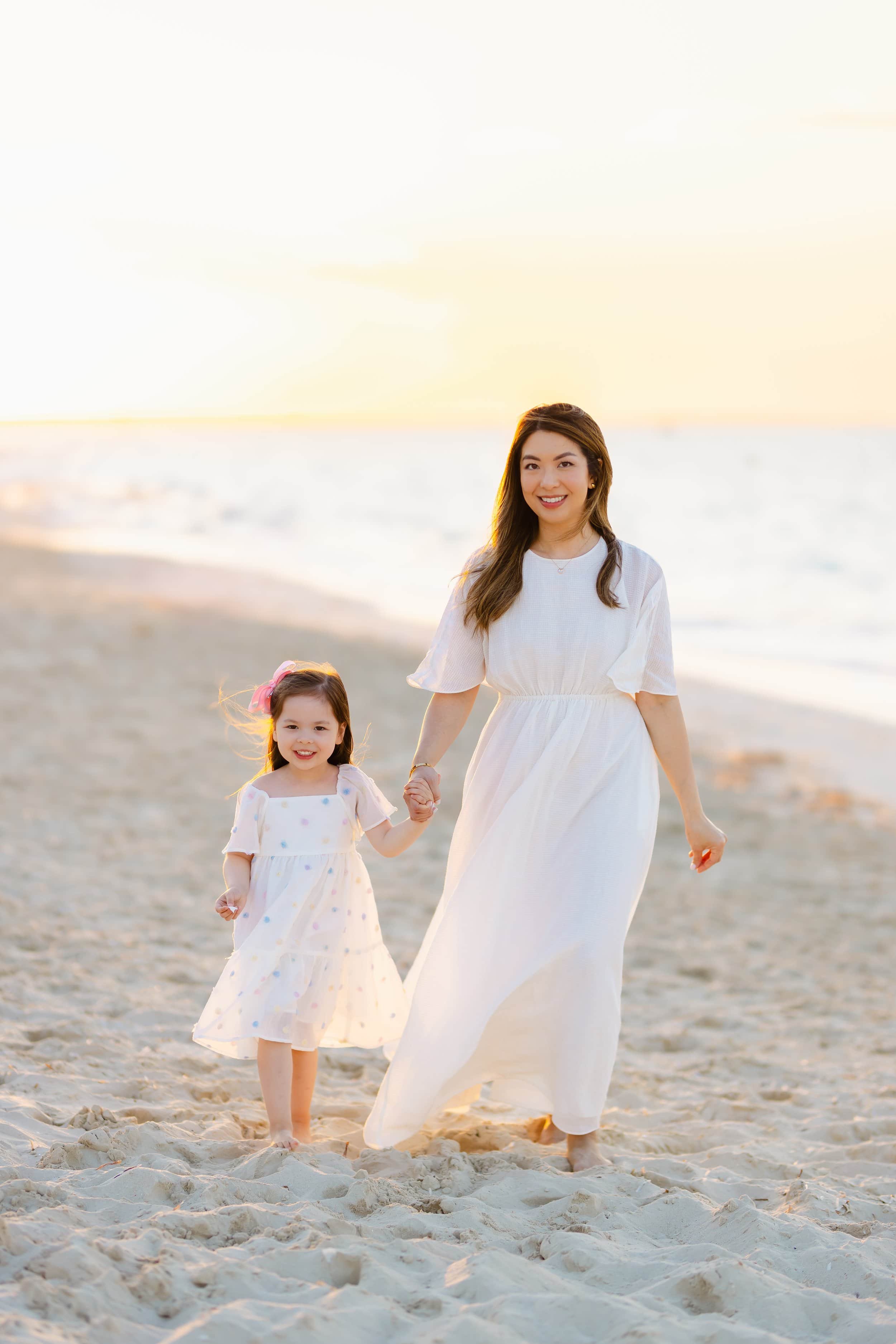 A woman and a young girl walking hand in hand on a beach during sunset, both wearing white dresses.