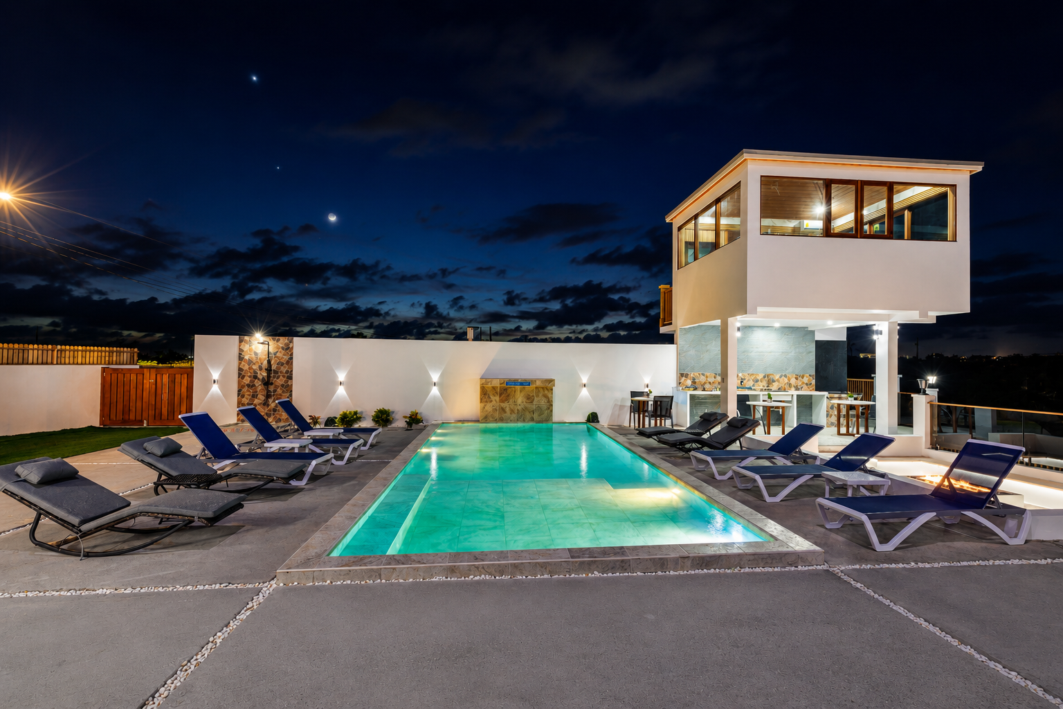 Night view of a modern backyard with a lit swimming pool, lounge chairs, and a two-story house with large windows, lit interior, and a cityscape in the distance.