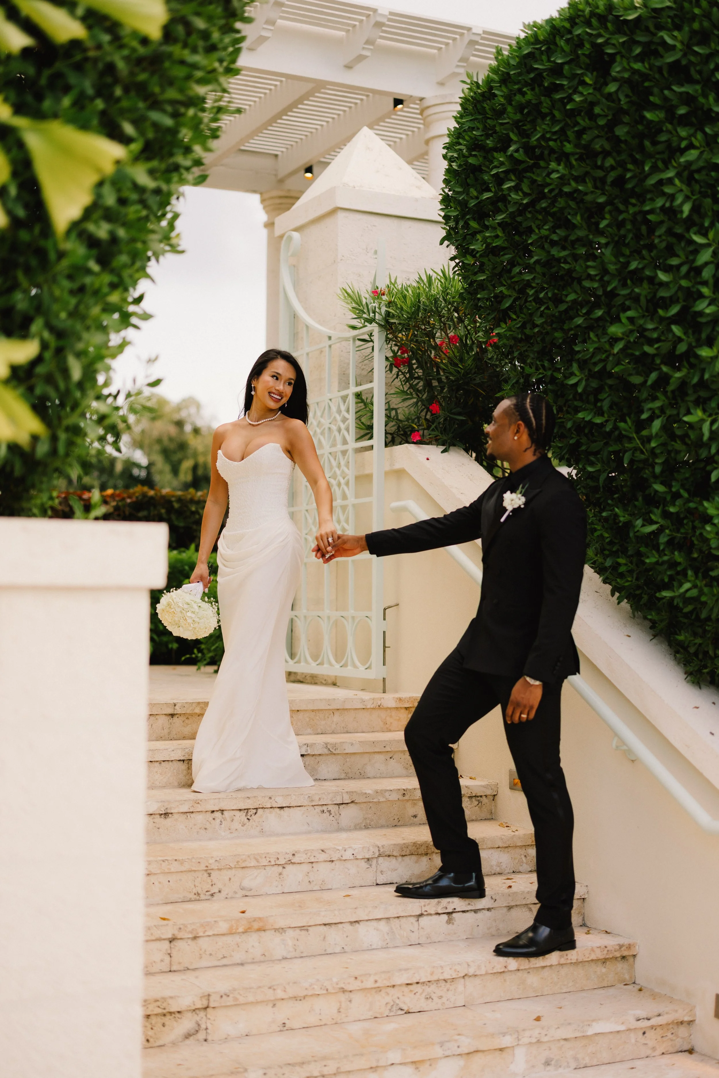 A bride and groom are holding hands on a staircase outside of a building, with greenery and flowers around them. The bride wears a strapless white gown and pearls, and the groom wears a black suit with a boutonniere.