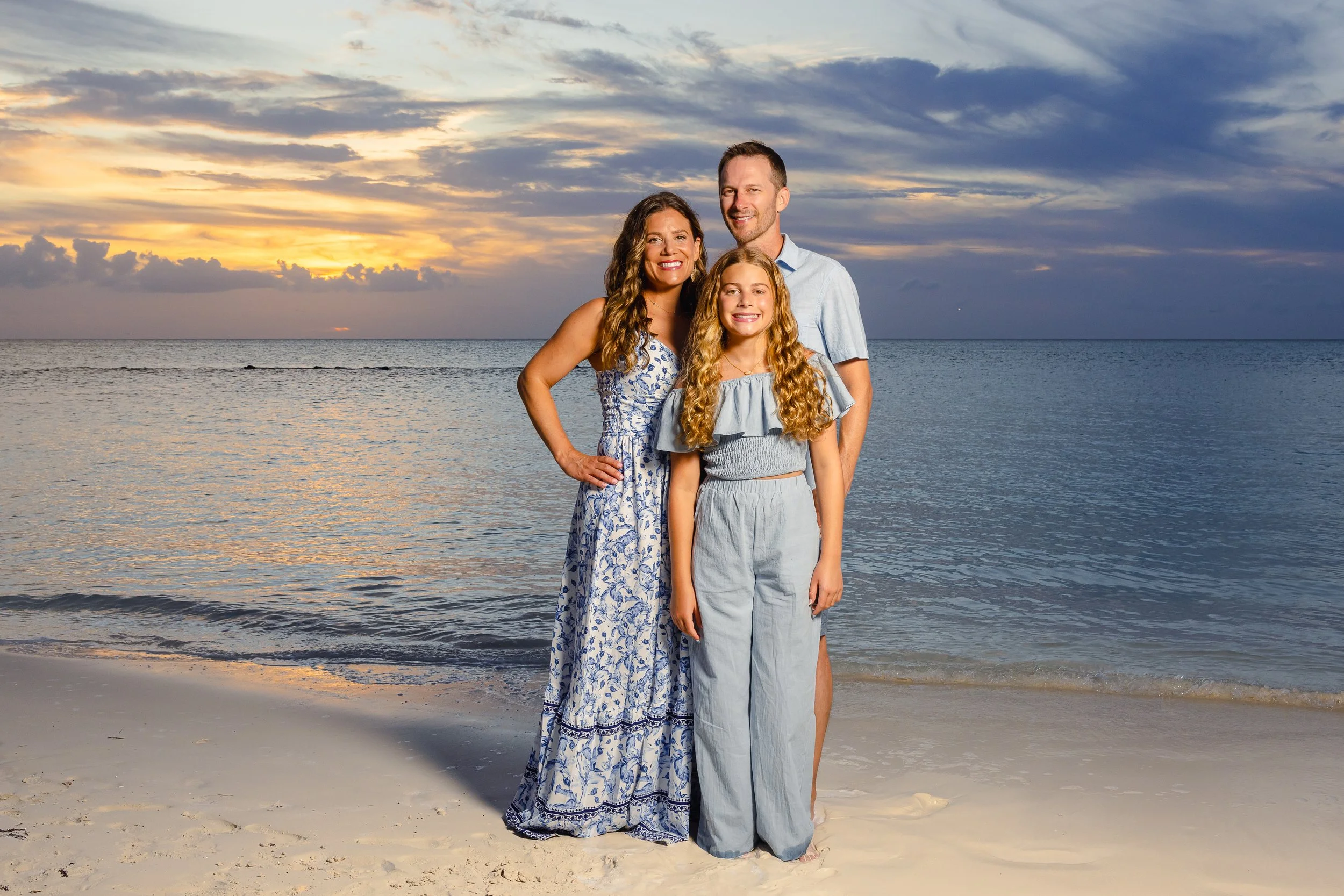 A family of three standing on a sandy beach at sunset, with the ocean and dramatic sky in the background. The woman is wearing a blue floral dress, the girl is in a light blue dress, and the man is in a light blue shirt and pants.