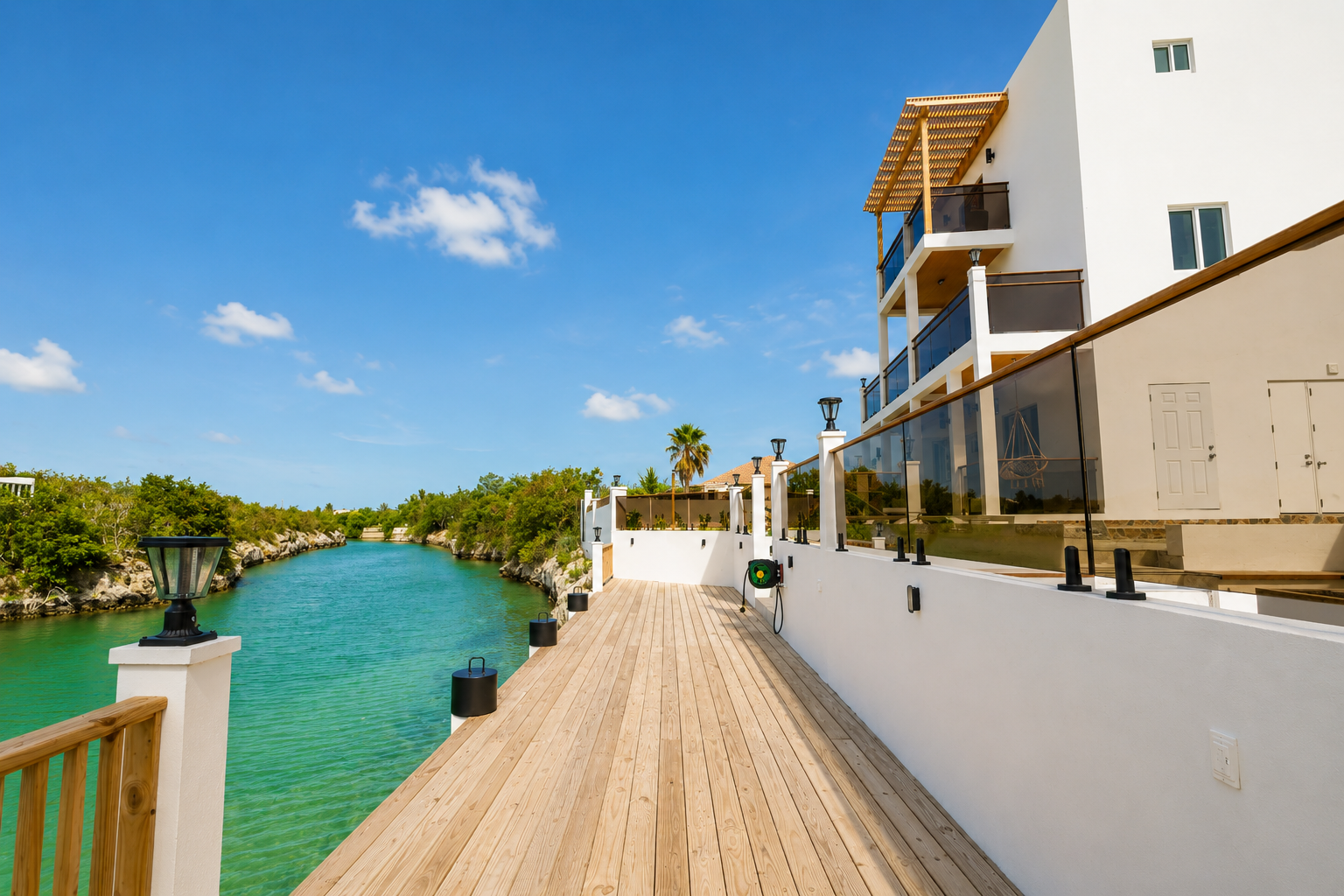 A wooden deck alongside a canal with clear blue water, white modern buildings with balconies, and green trees under a bright blue sky with scattered clouds.