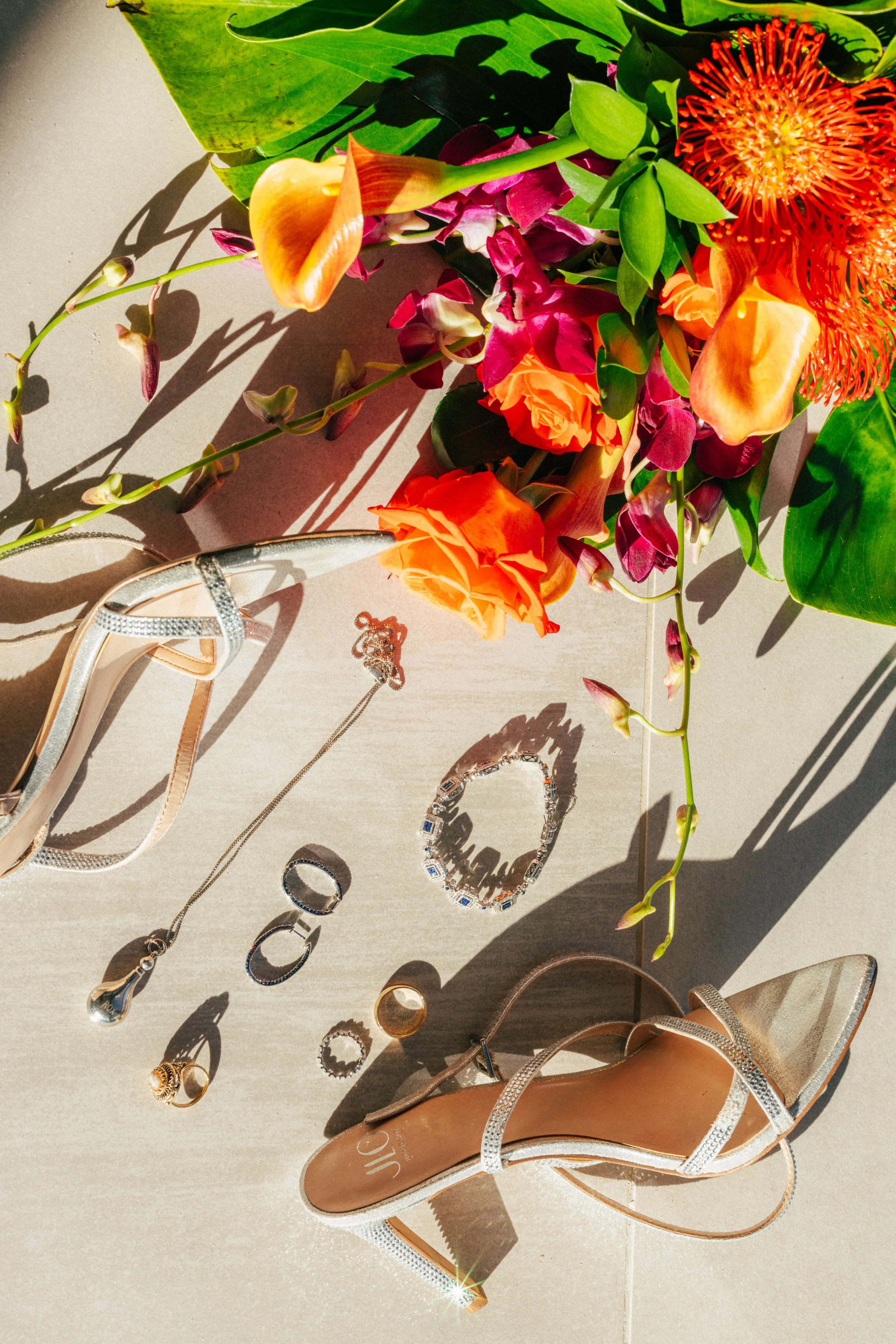 A flat lay of assorted jewelry including rings, earrings, a bracelet, and necklaces, alongside a beige sandal with rhinestones on a light-colored surface, with a bouquet of pink, orange, and red flowers and green leaves.