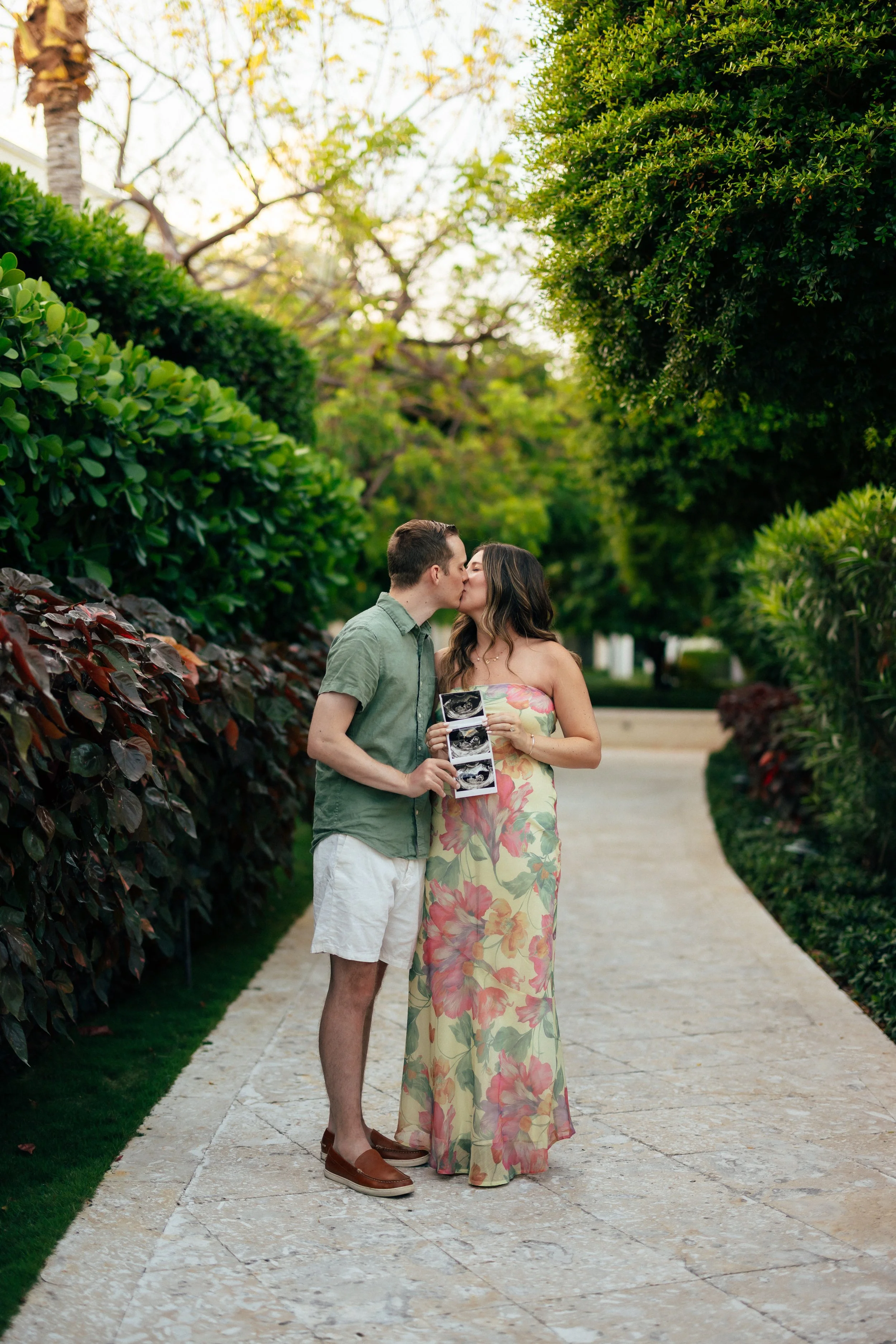 A young couple sharing a kiss in a park, the woman holding ultrasound images, surrounded by lush green trees and bushes on a sunny day.