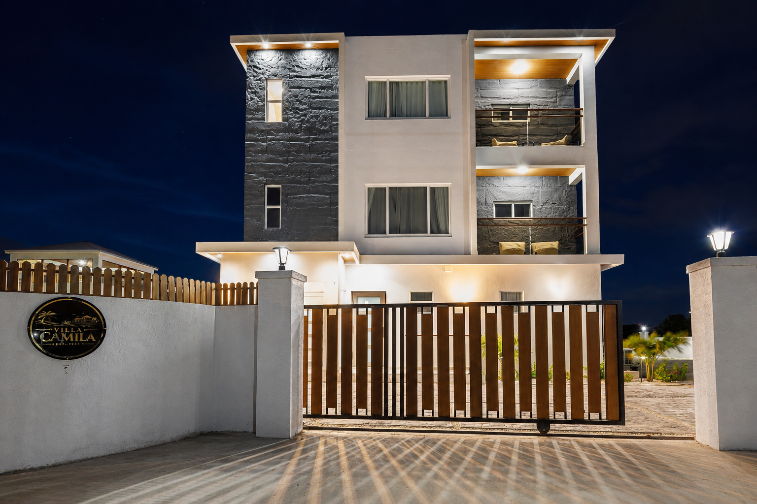 Night view of a modern, multi-story residential building with white and grey exterior walls, illuminated by outdoor lighting, surrounded by a white concrete fence with a wooden gate, and a sign that reads "VILLA CAMILA."