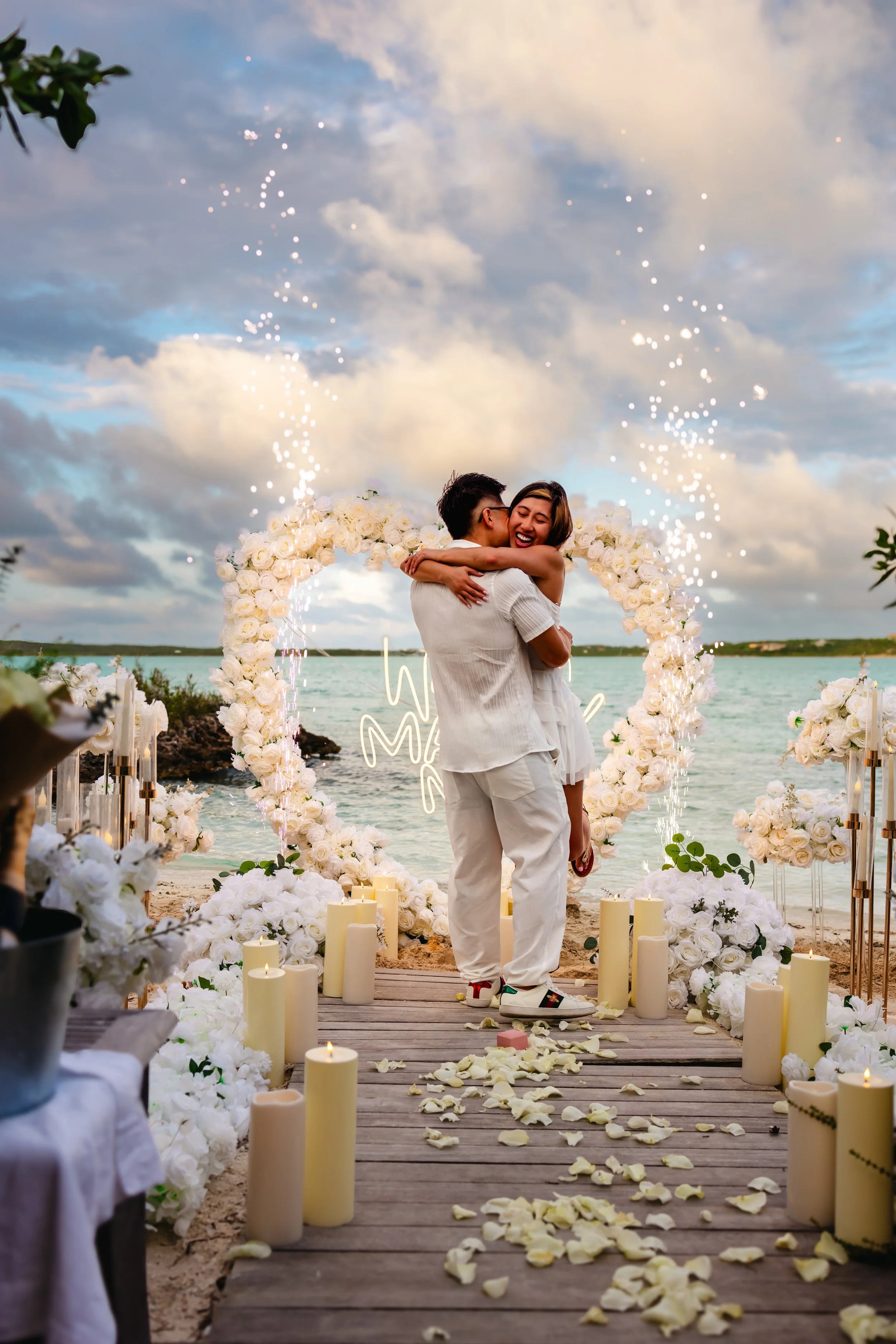 A couple joyfully hugging during a wedding ceremony by the water, with floral decorations, candles, and rose petals on a wooden pathway under a cloudy sky.