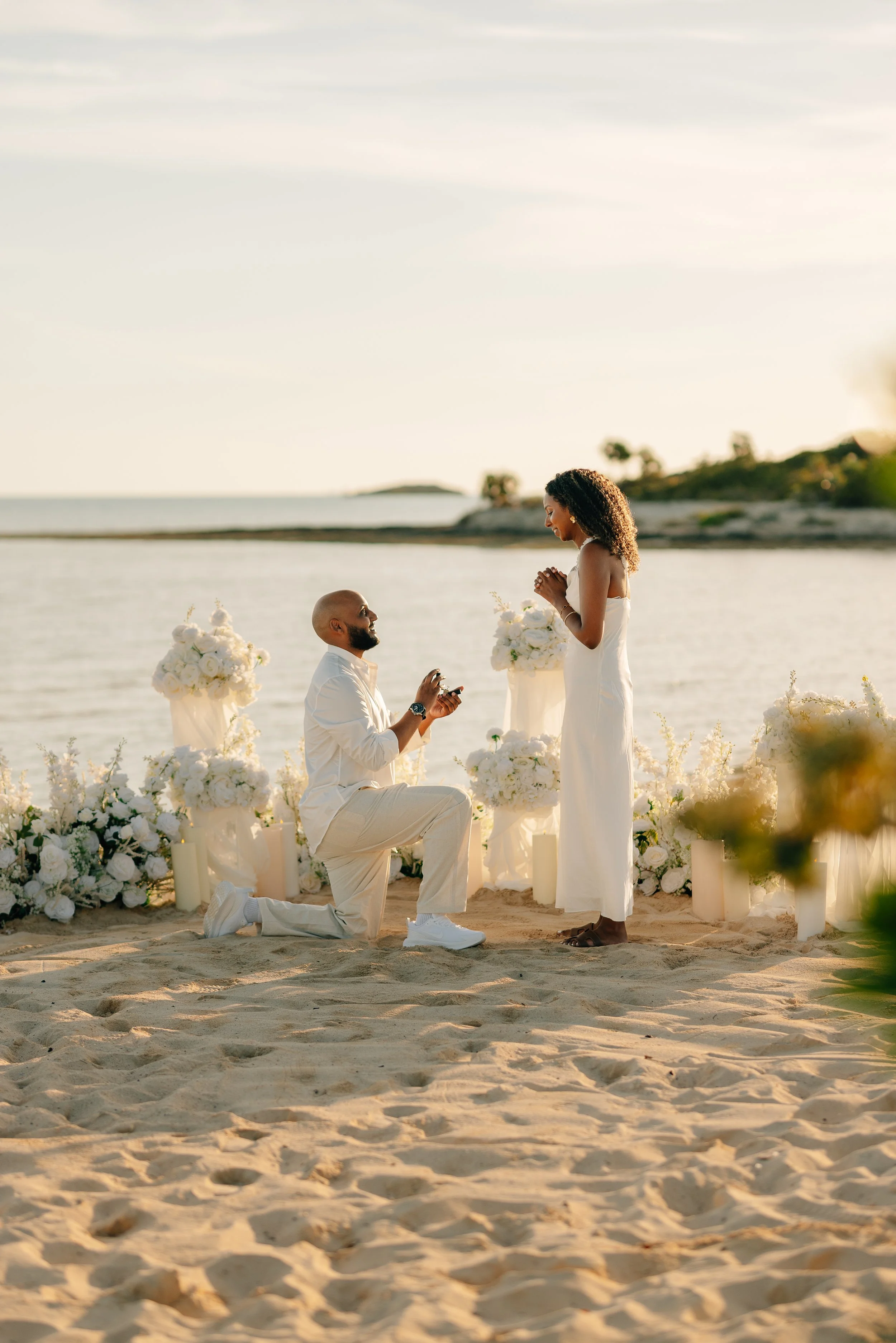 A man proposes marriage to a woman on a beach during sunset, with flowers and candles arranged around them.