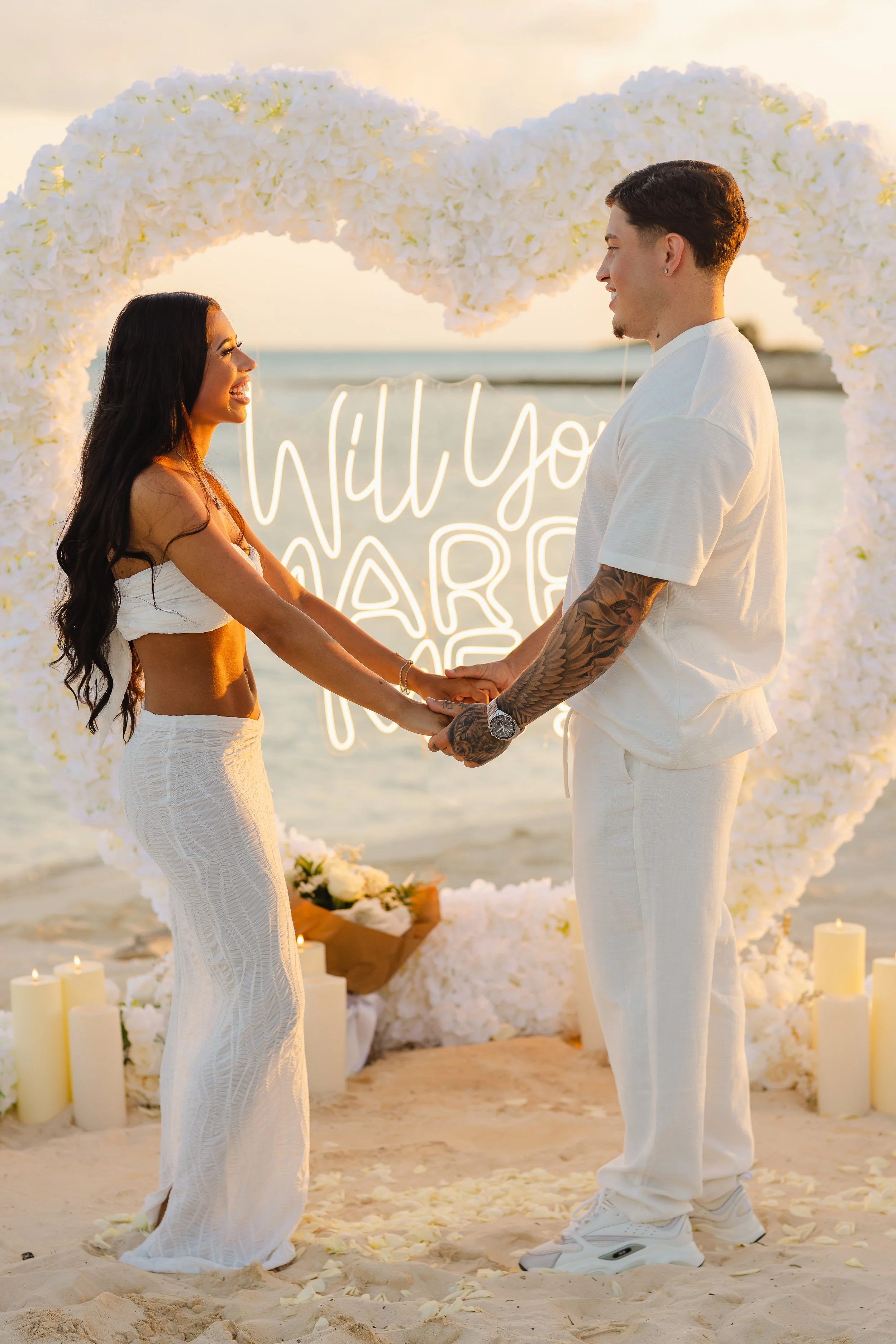 A couple getting married on the beach during sunset, standing face to face holding hands, with a heart-shaped floral arch and a neon sign that reads 'Will you marry me' behind them.