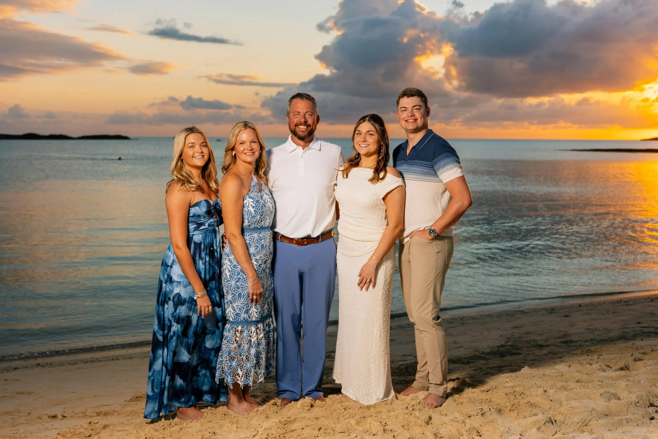 A group of five people standing on a beach during sunset. The group includes three women and two men, smiling and wearing casual or semi-formal attire. The background features a calm ocean, a cloudy sky with warm sunset colors, and a distant landmass