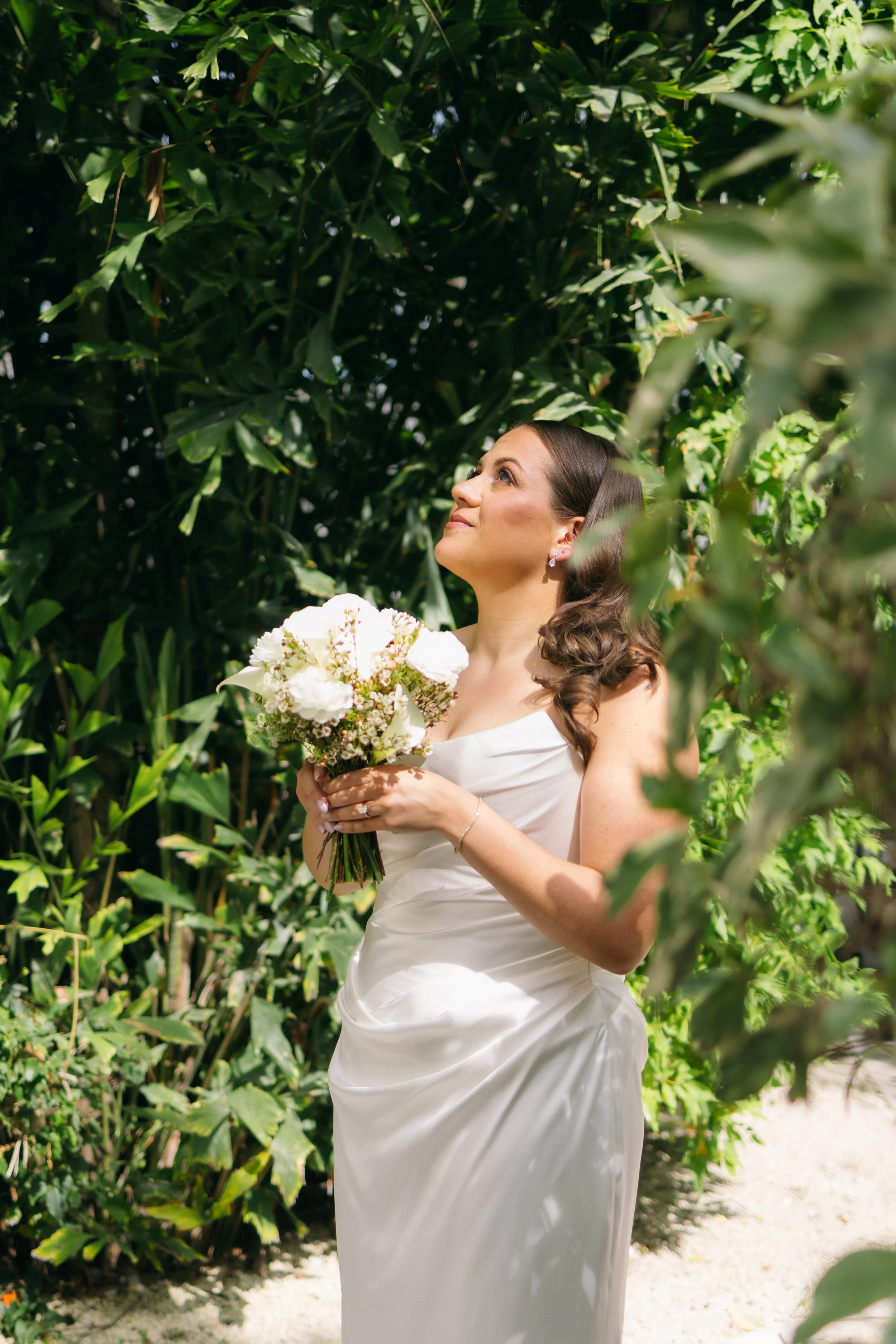 A bride in a white wedding dress holding a bouquet of flowers standing outdoors in front of green foliage, looking up.