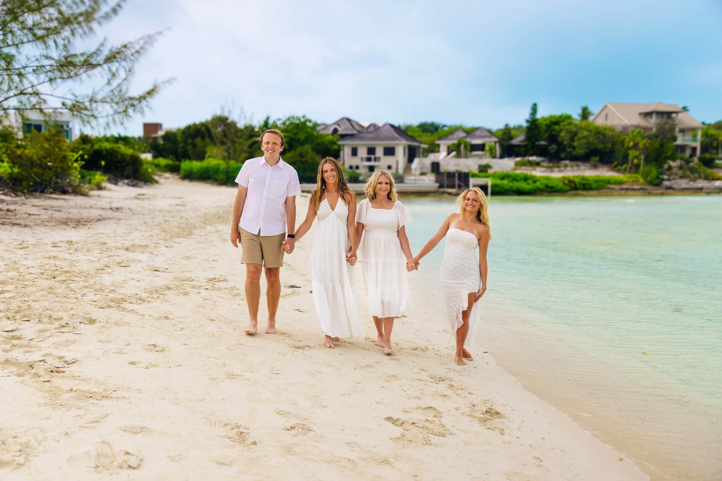 Four family members walking along a sandy beach holding hands, with houses and greenery in the background.