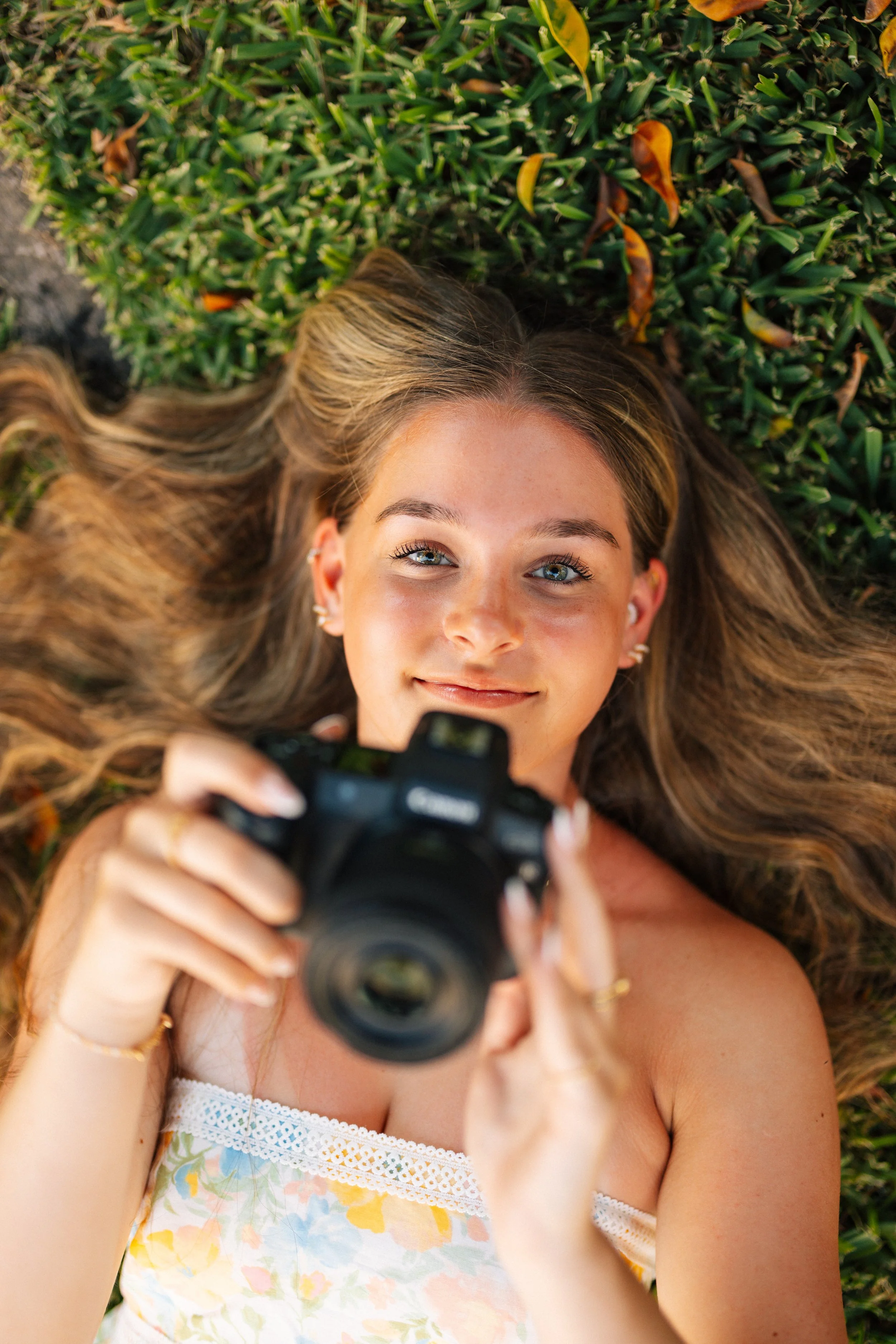 Young woman with long hair lying on grass, taking a selfie with a camera, looking up at the camera with a smile.