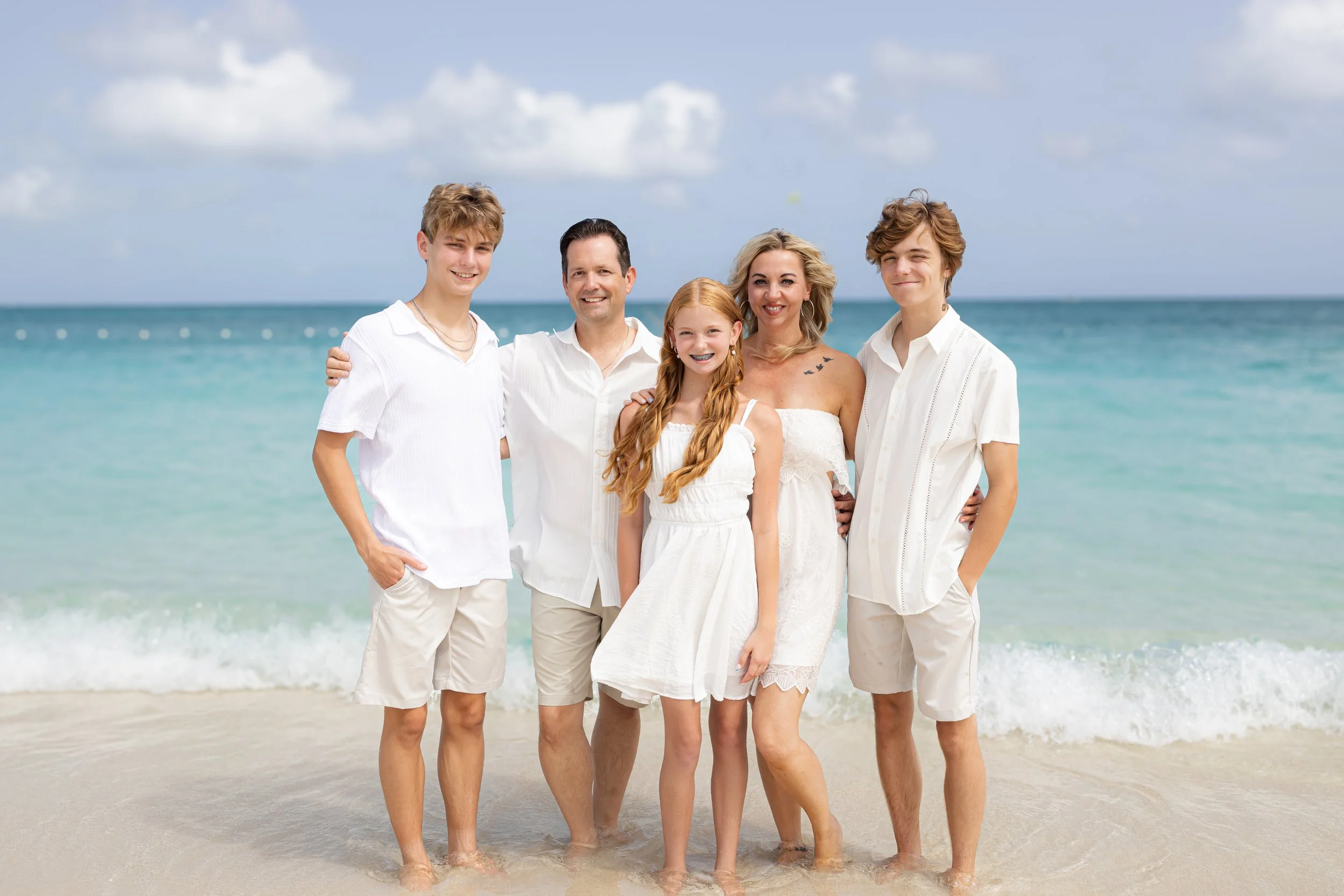 A family of six people standing close together on the beach near the water, all dressed in white summer clothes, smiling at the camera.