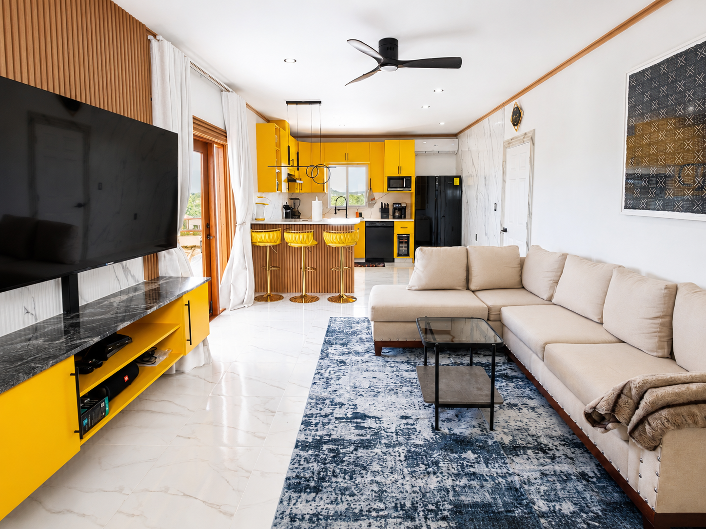 Living room with beige sectional sofa, blue patterned rug, black coffee table, wall-mounted TV on yellow and black stand, open kitchen with yellow cabinets, bar stools, and sliding glass doors with white curtains.