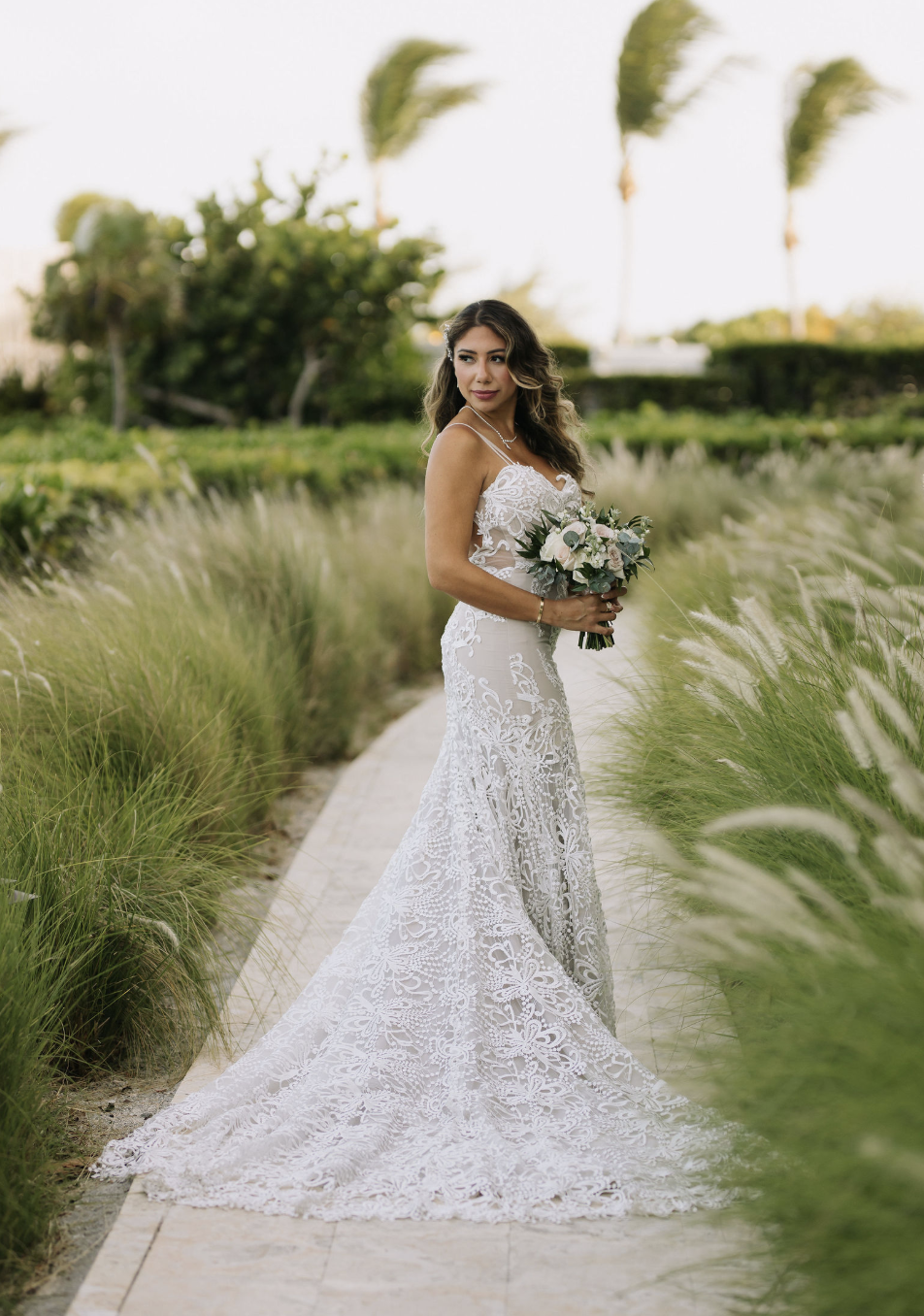 A bride in a white lace wedding dress holds a bouquet of flowers, standing on a stone pathway in a green outdoor setting with palm trees in the background.