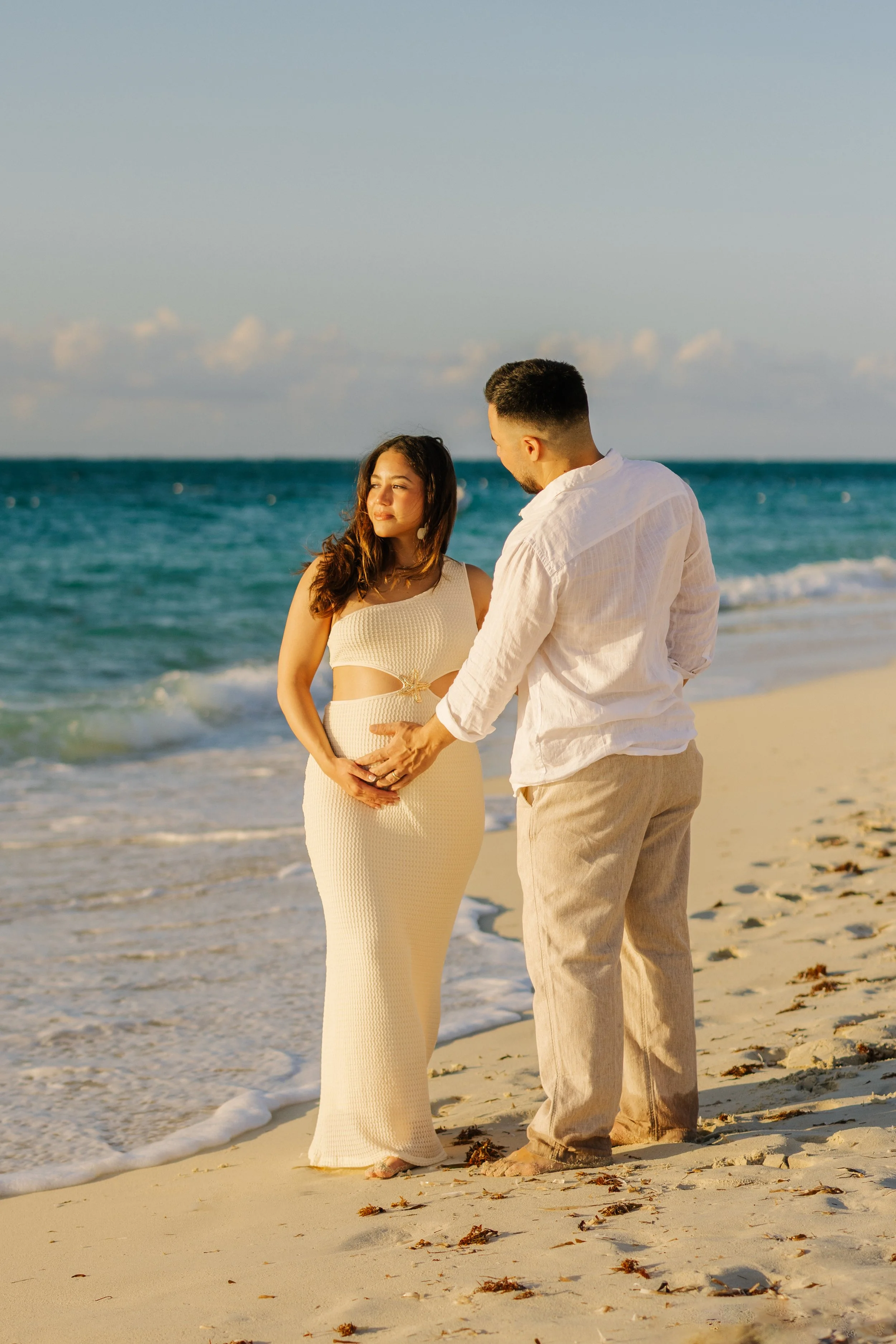 A pregnant woman and a man stand on the beach facing each other, holding hands and looking into each other's eyes, with the ocean and a partly cloudy sky in the background.