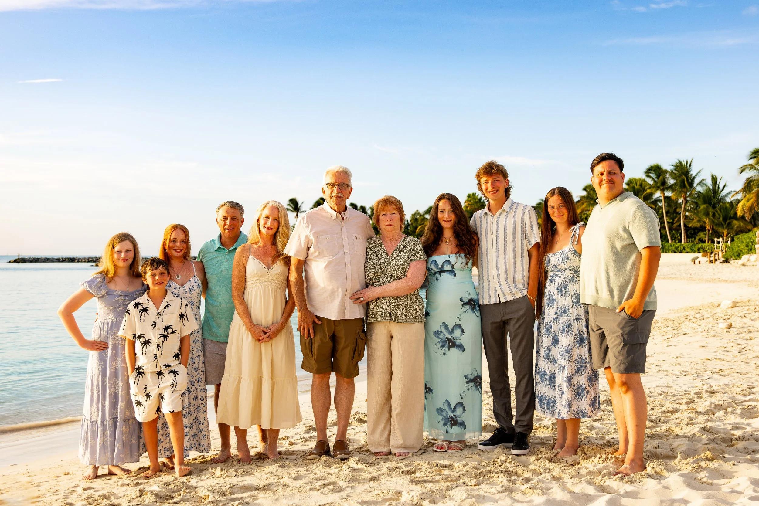 A multigenerational family of fourteen standing on a beach, smiling, with palm trees and the ocean in the background during the daytime.