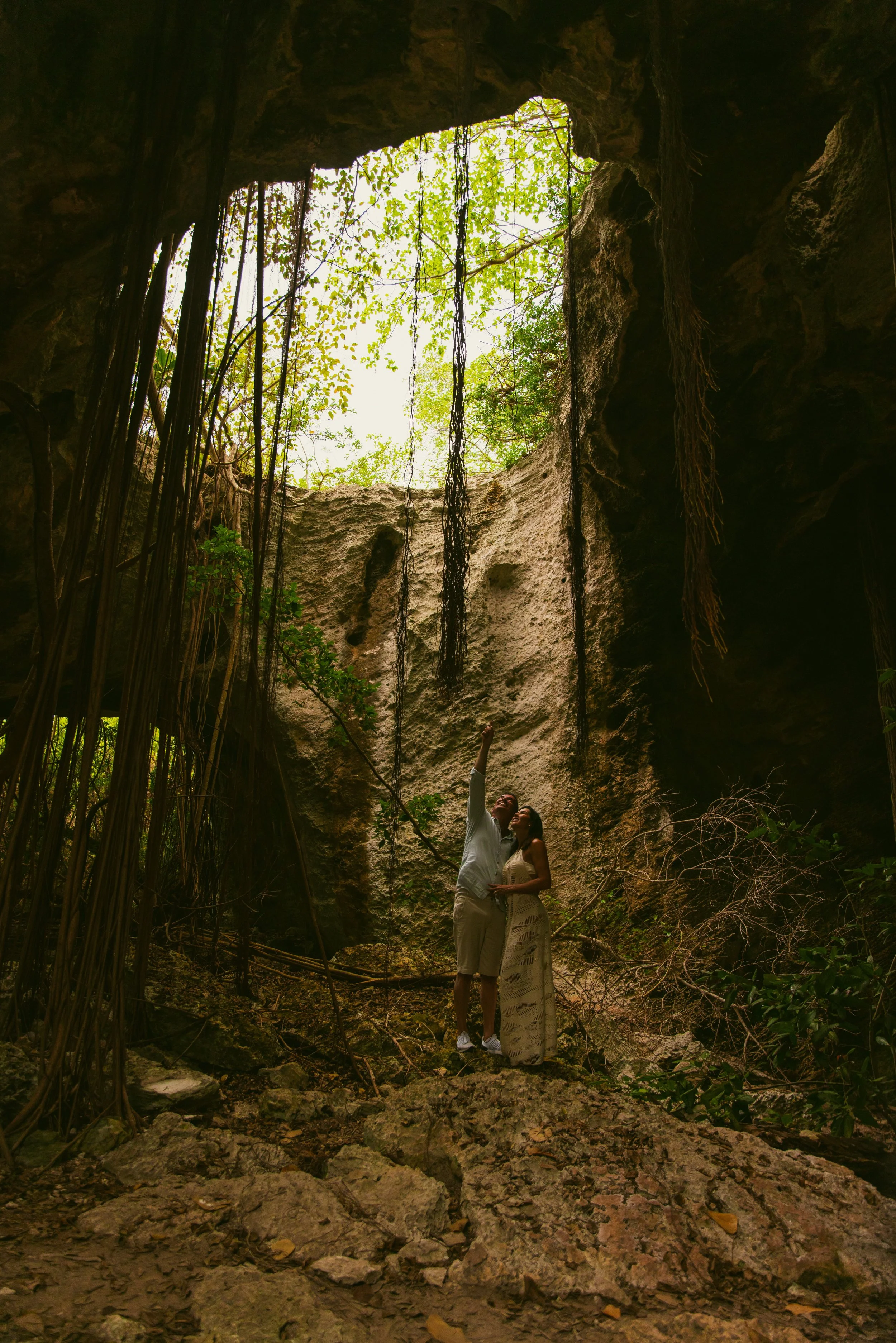 A couple standing inside a large cave, looking up at the sky through an opening with green trees visible above. The ground is rocky, and vines hang from the cave ceiling.