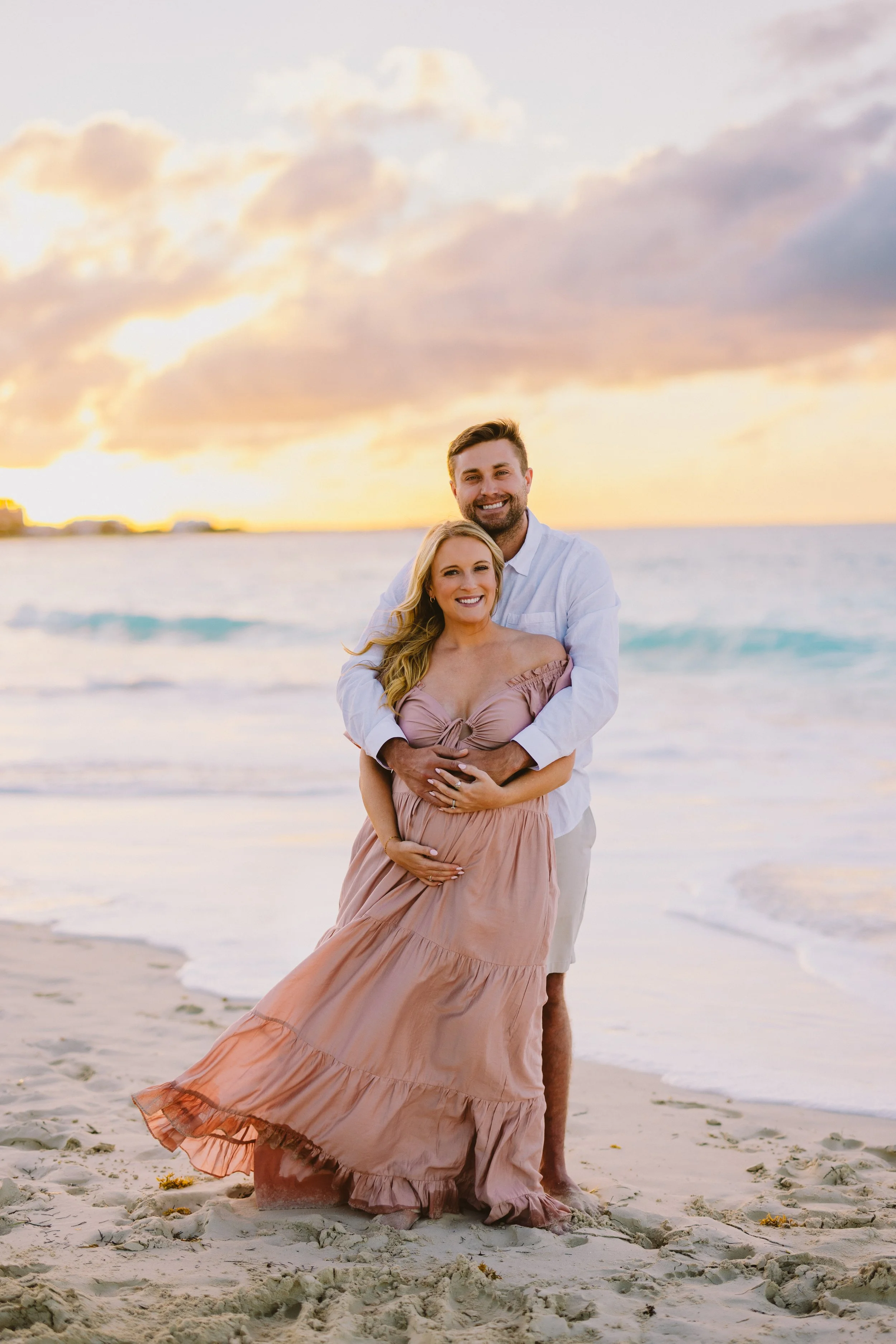 A couple dressed in light clothing on a beach at sunset, with the woman holding her pregnant belly and the man embracing her from behind.