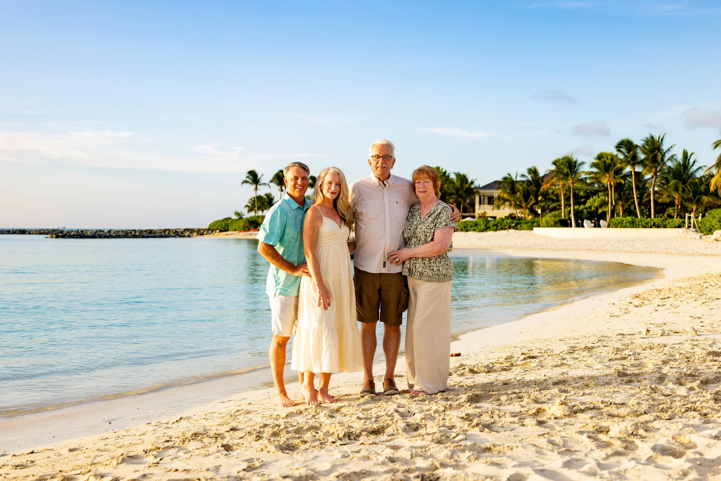 Four family members standing arm-in-arm on a sandy beach with palm trees and a house in the background, enjoying a sunny vacation by the ocean.