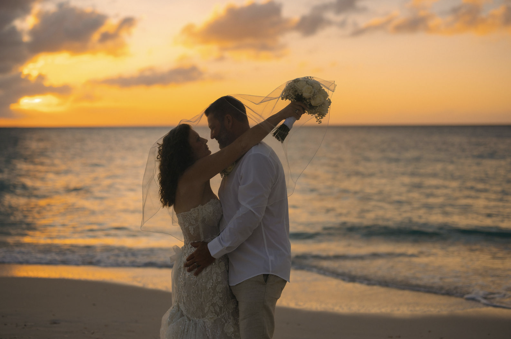 A newlywed couple stands close together on the beach during sunset, with the bride holding a bouquet of white flowers and a sheer veil over her hair, as the groom looks down at her, with the ocean and a colorful sky in the background.