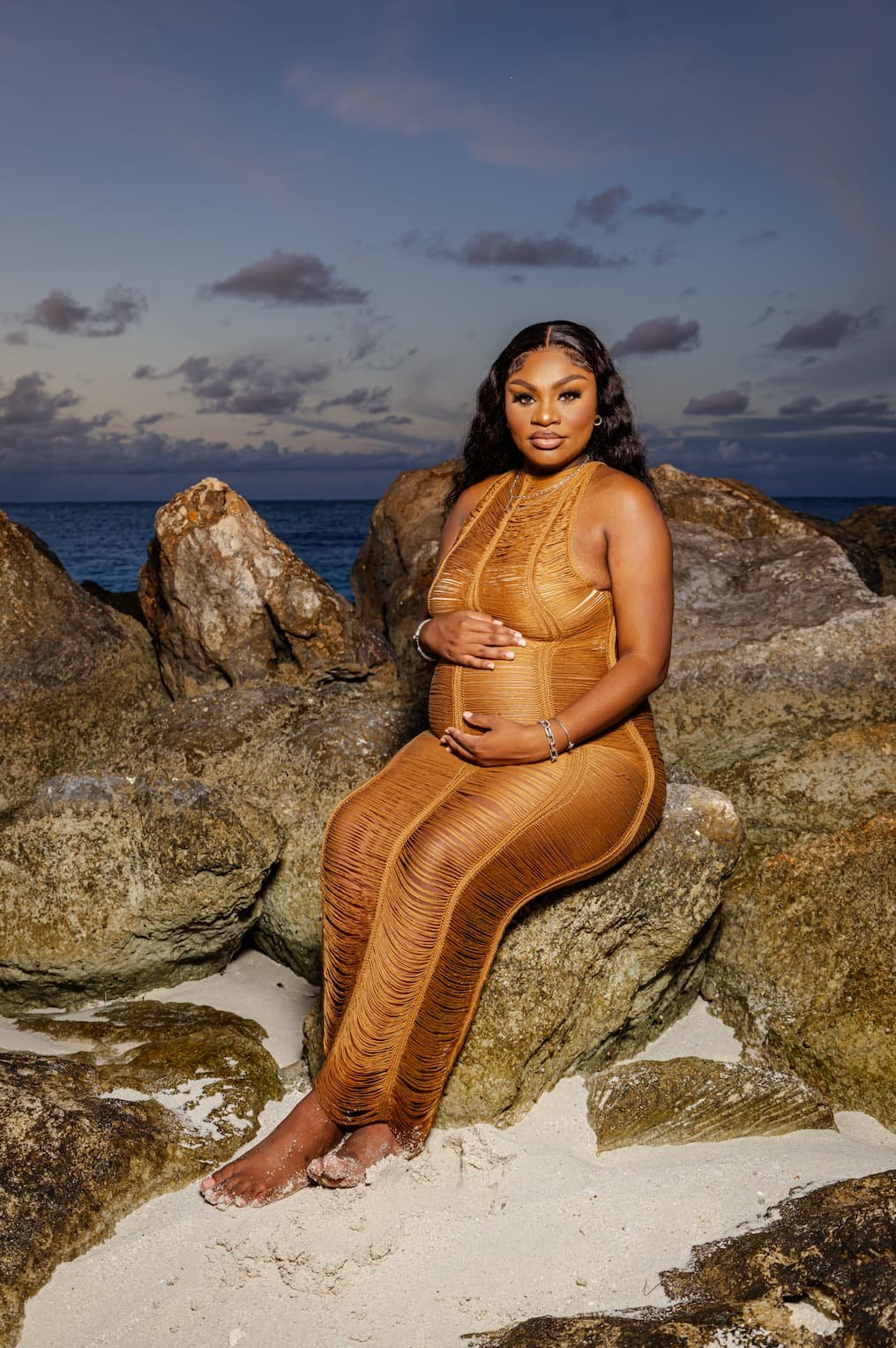 A woman in an orange dress sitting on rocks by the beach with the ocean and cloudy sky in the background.