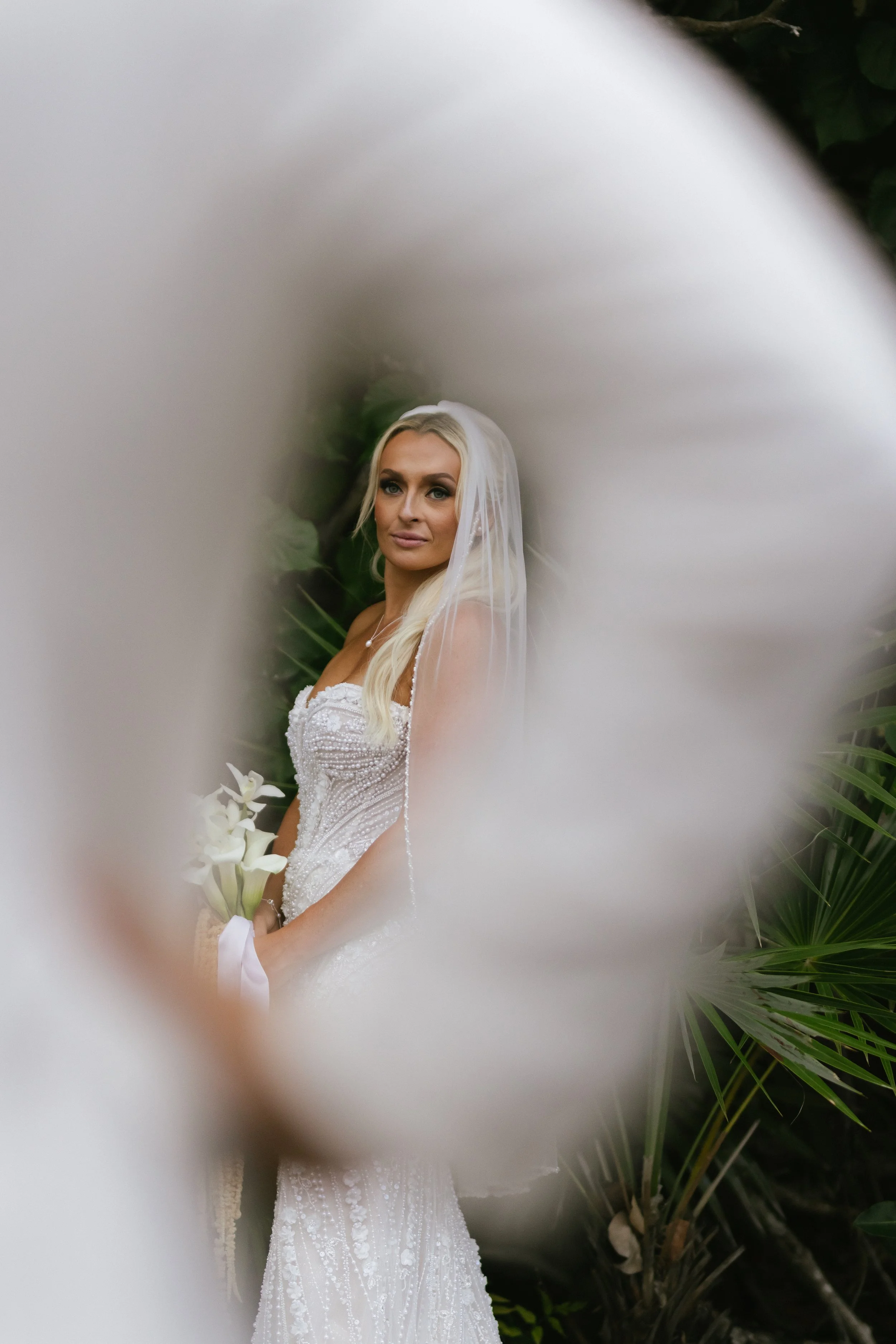 A bride with blond hair and blue eyes wearing a white wedding gown and veil, holding a bouquet of white calla lilies, standing outdoors near green plants.