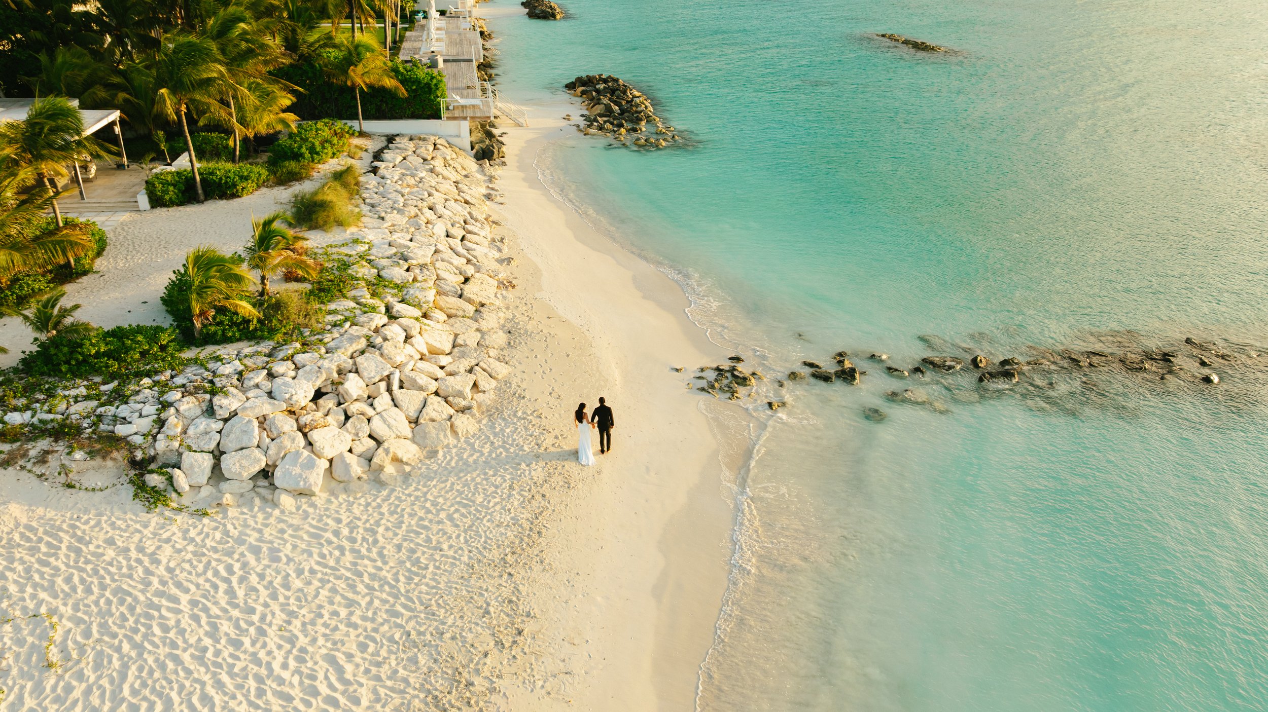An aerial view of a couple walking on a sandy beach near turquoise water, with greenery and rocks along the shore, and some buildings and palm trees above.