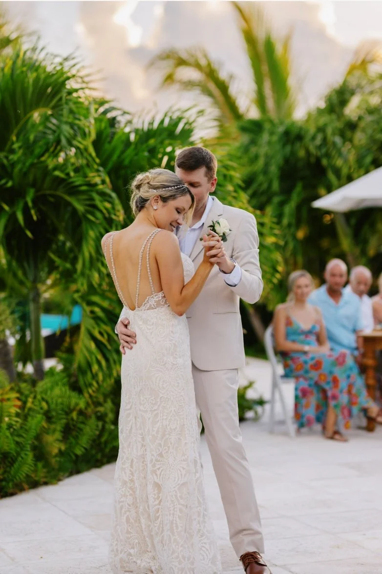 Bride and groom dancing at outdoor wedding reception surrounded by greenery, with guests seated in the background.