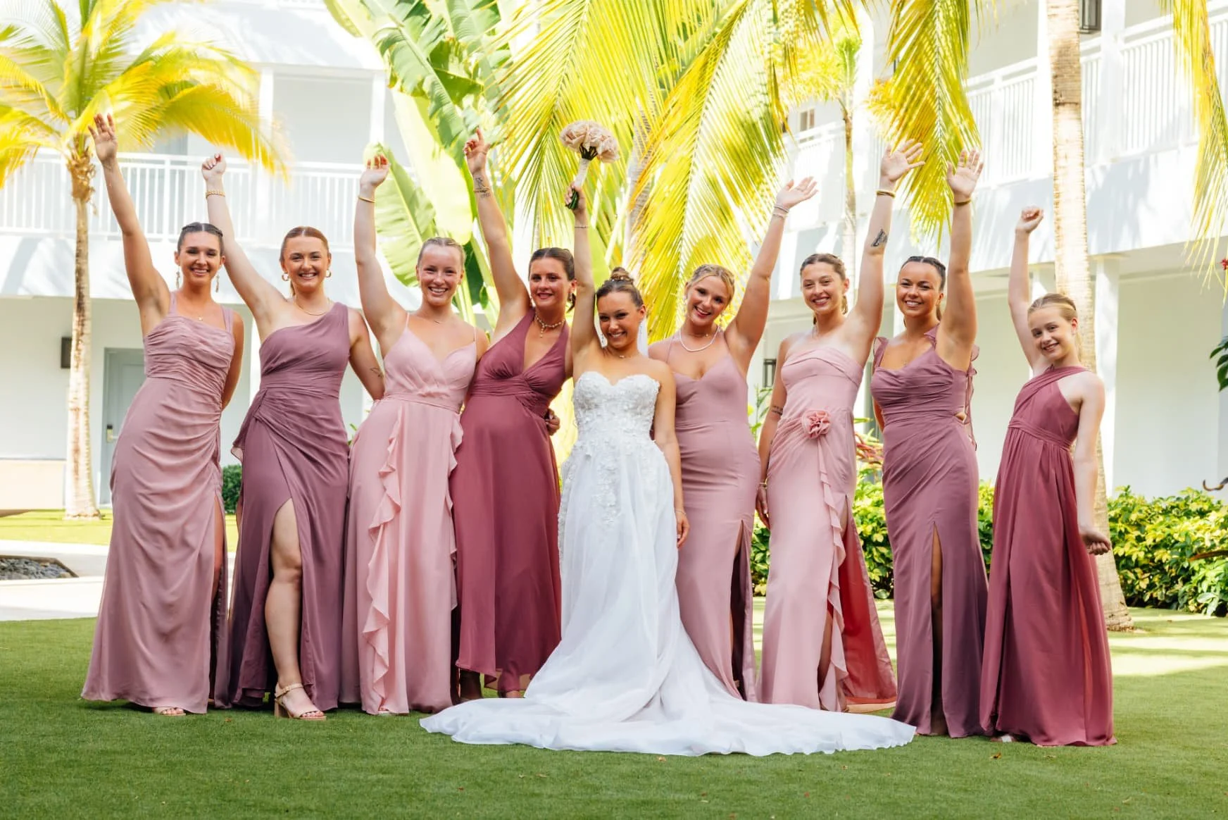A group of women, likely bridesmaids, standing outdoors on green grass with palm trees and a white building in the background. They are wearing various shades of pink and purple dresses. The woman in the center is dressed in a white wedding gown and holding a bouquet. They are smiling, cheering, and raising their hands in celebration.