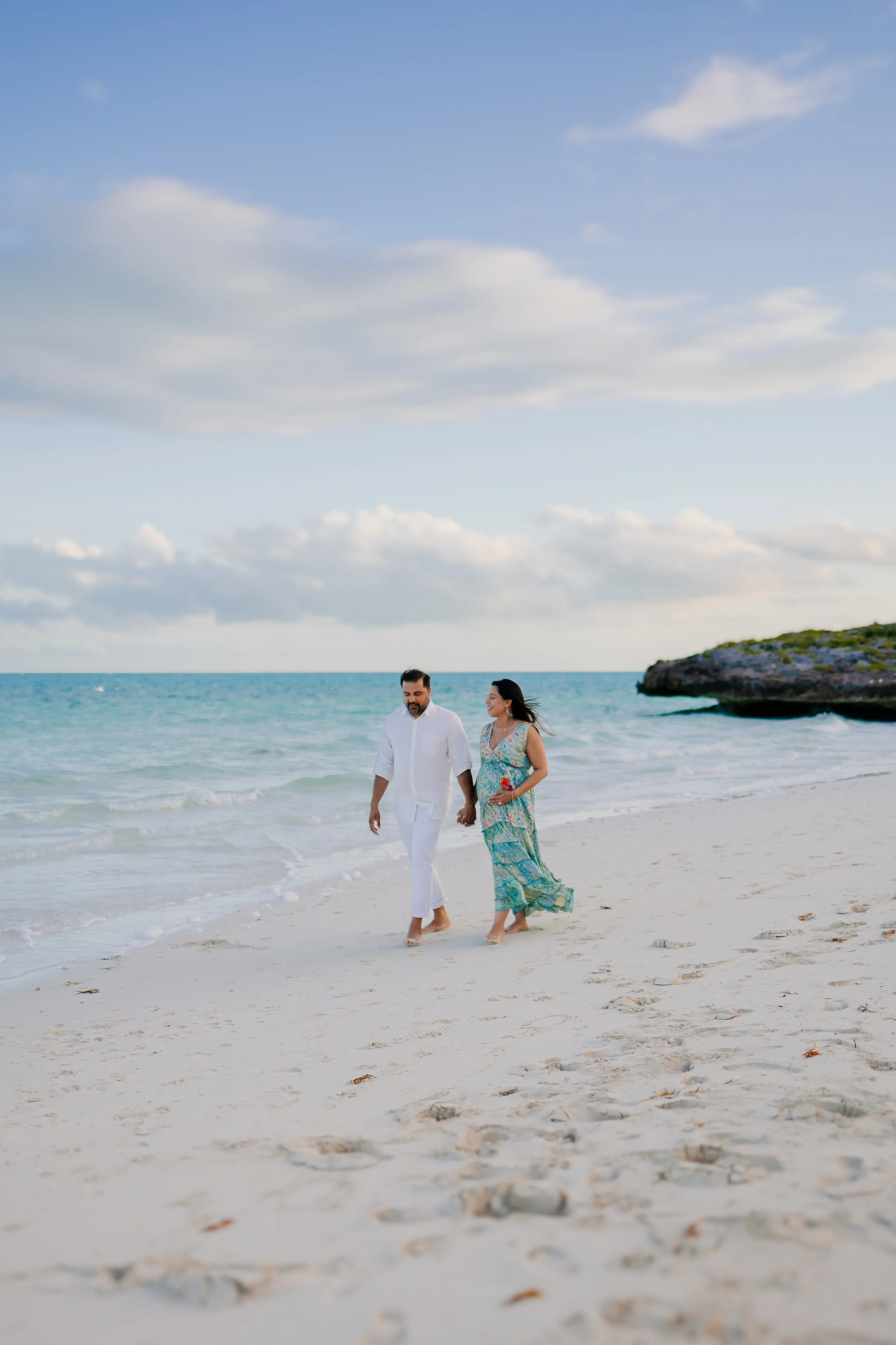 A couple walking along the beach holding hands, with ocean waves and a cloudy sky in the background.