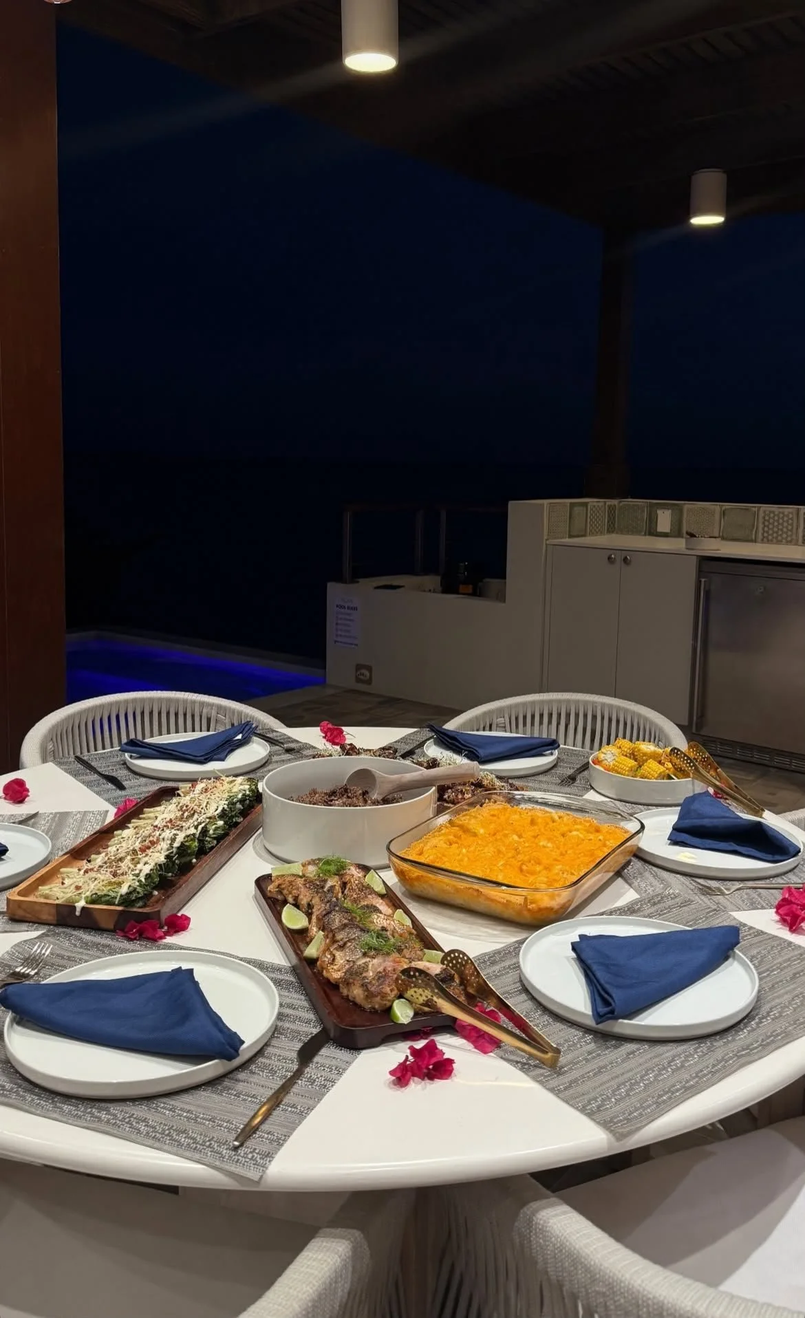 Dinner table set outdoors with various dishes including grilled fish, baked casserole, and salads. Blue napkins at each place setting, surrounded by chairs, with a dark night sky and outdoor kitchen area in the background.