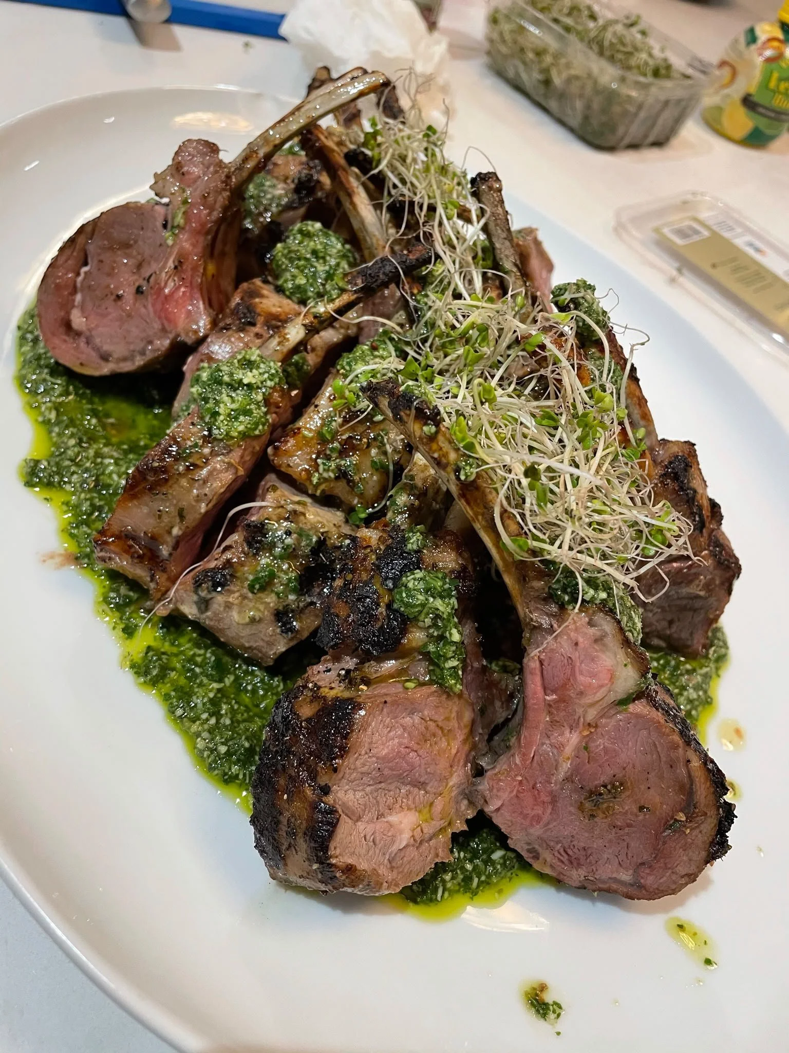 Close-up of a plate of cooked, seasoned lamb chops garnished with green herbs and microgreens, with green sauce drizzled on the meat, on a white plate.