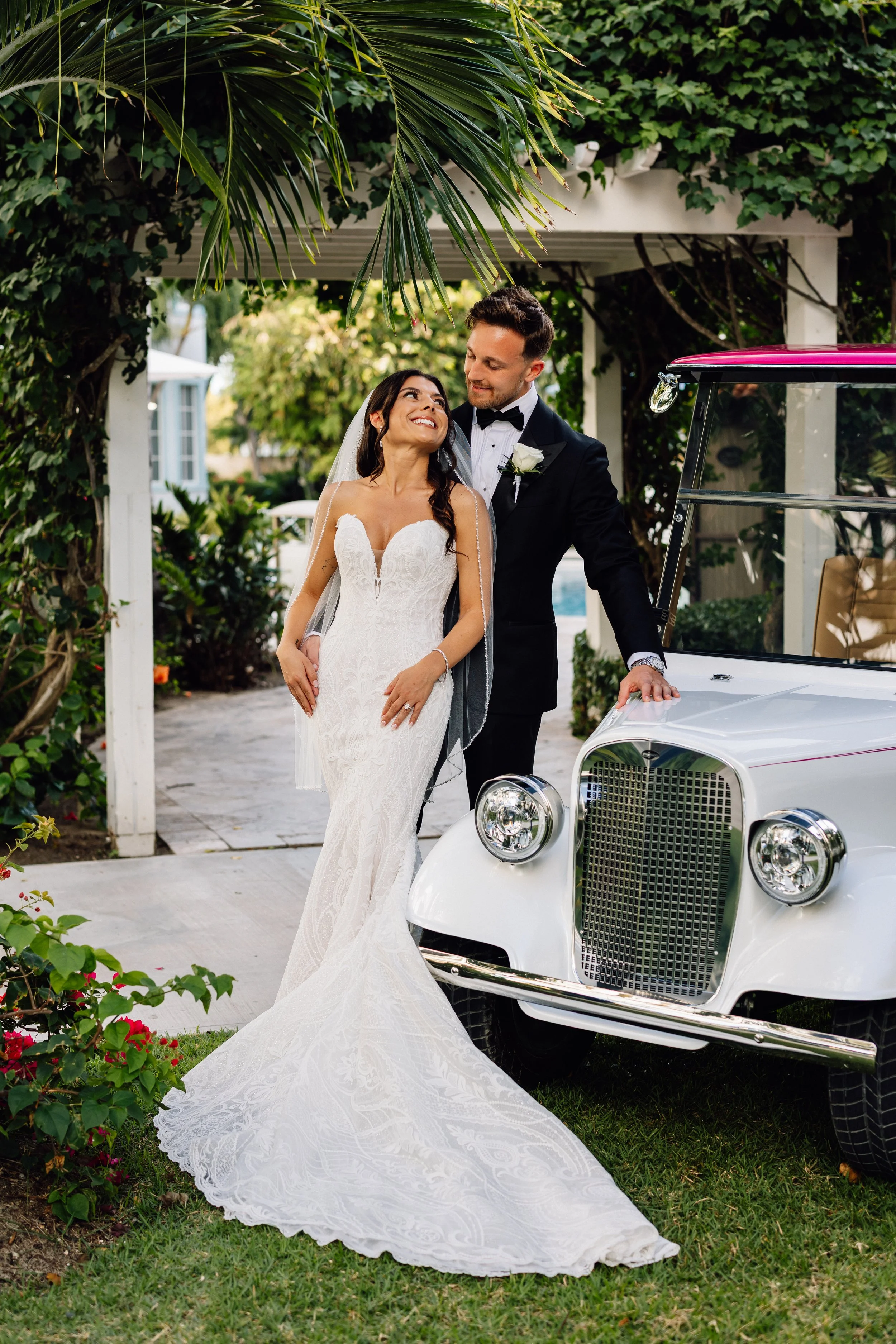 A bride in a white lace wedding dress and a groom in a black tuxedo stand together near a white vintage car, smiling at each other under a canopy with lush greenery and flowers.