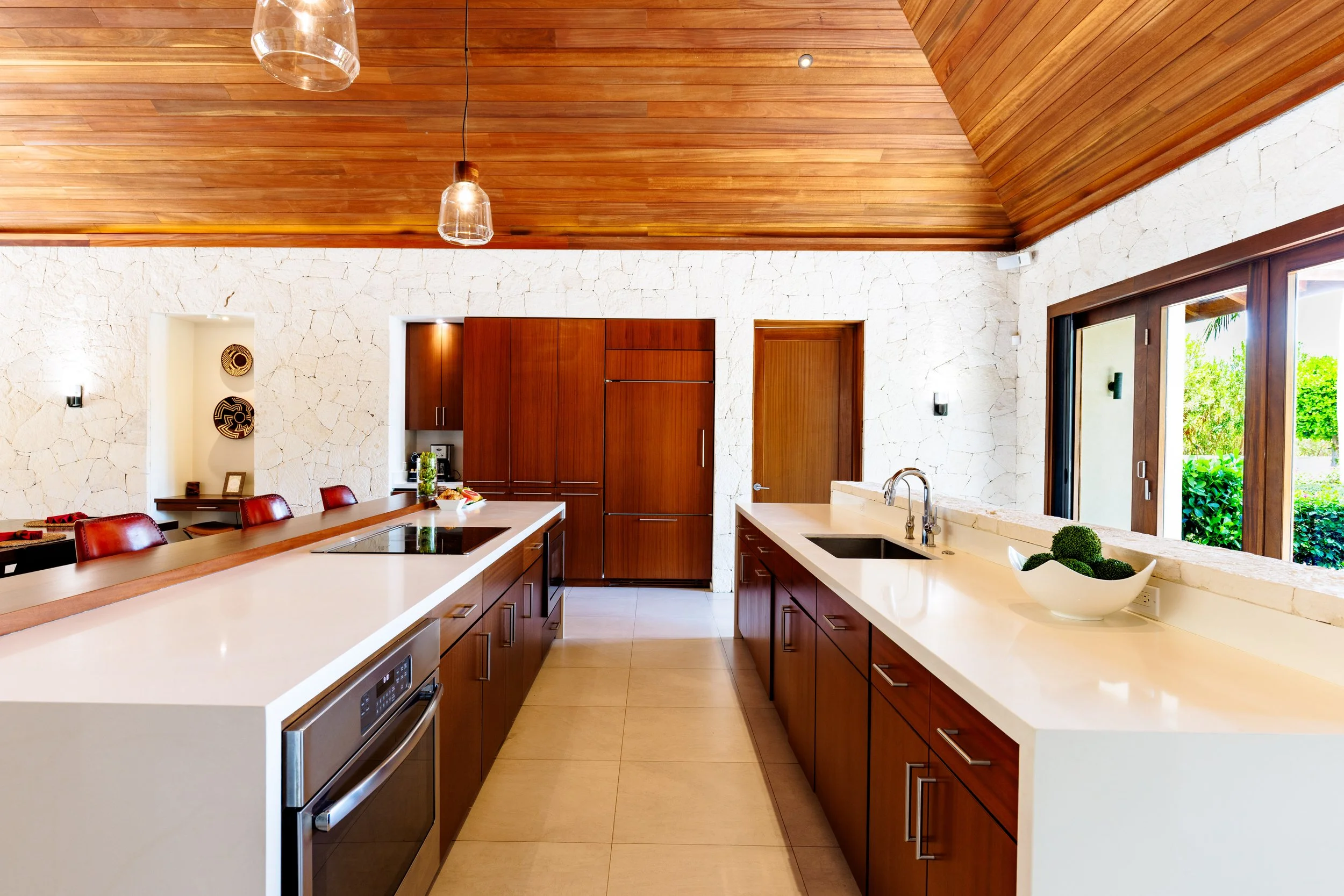 Modern kitchen with white countertops, wooden cabinets, a large sink, and a view of outdoors through sliding glass doors.