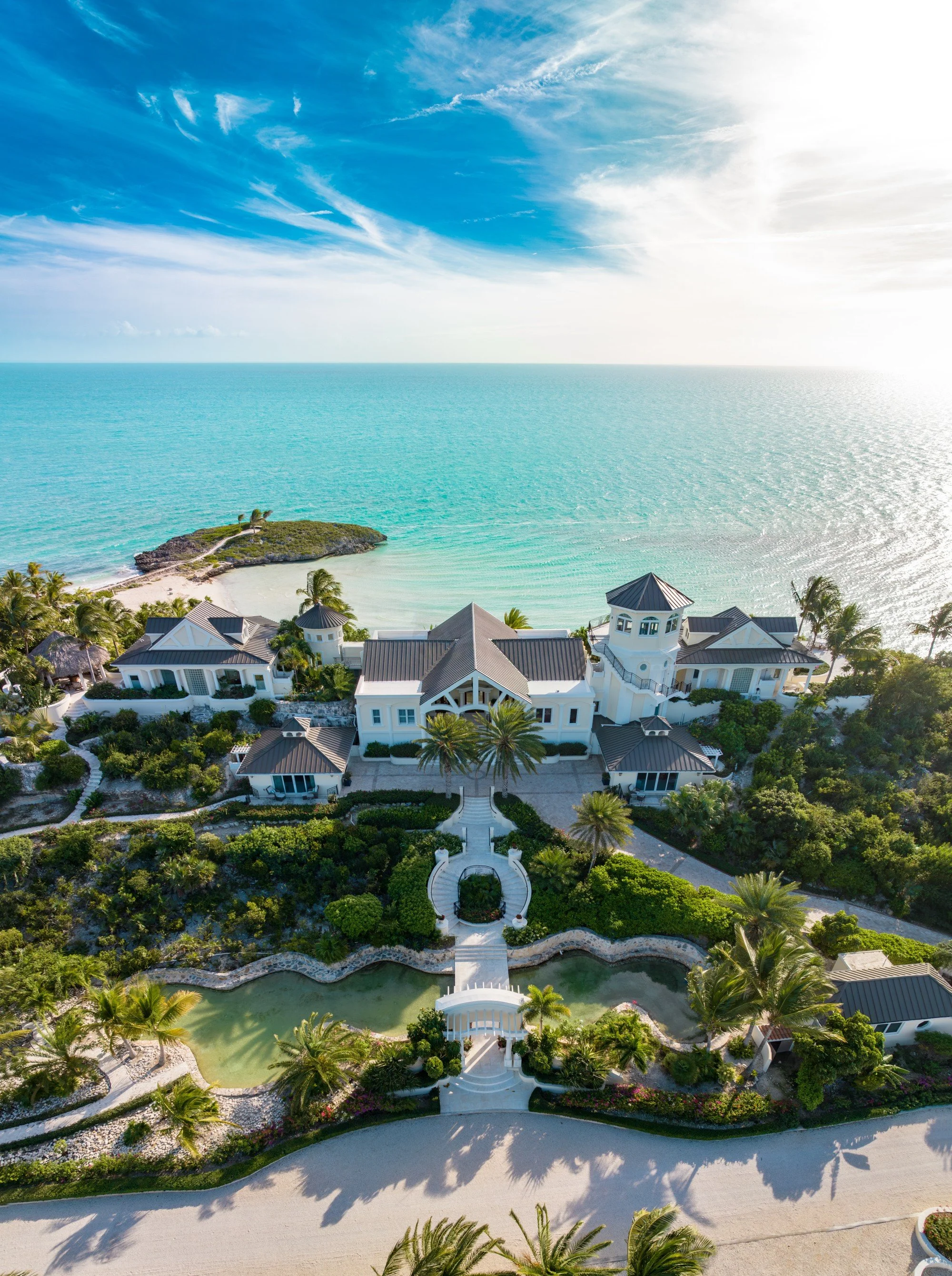 Aerial view of a luxurious seaside estate with white buildings, a manicured garden, a winding pathway, a swimming pool, and clear turquoise ocean water in the background under a blue sky with wispy clouds.