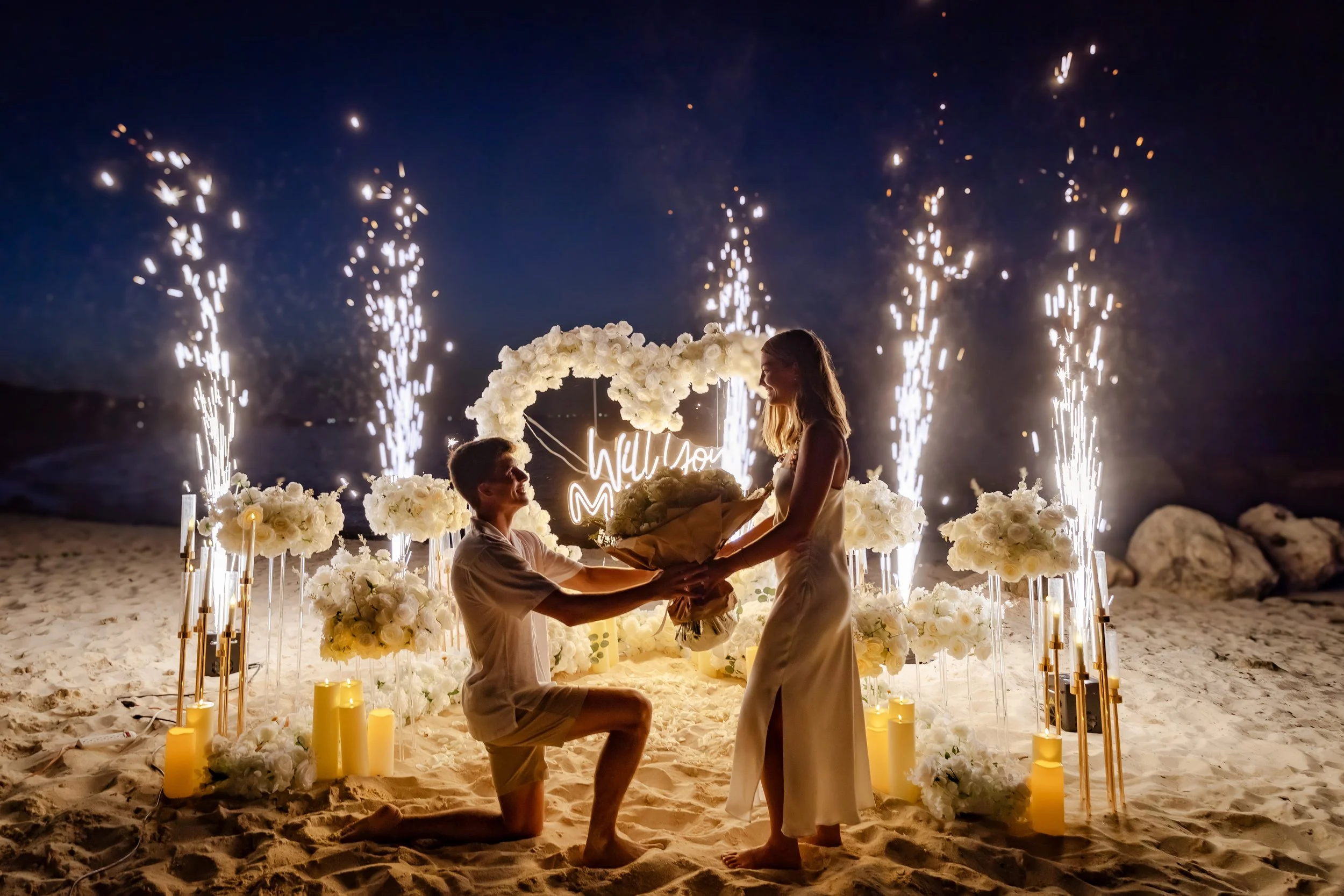 A couple getting engaged on a decorated beach at night with fireworks and floral decorations surrounding them.
