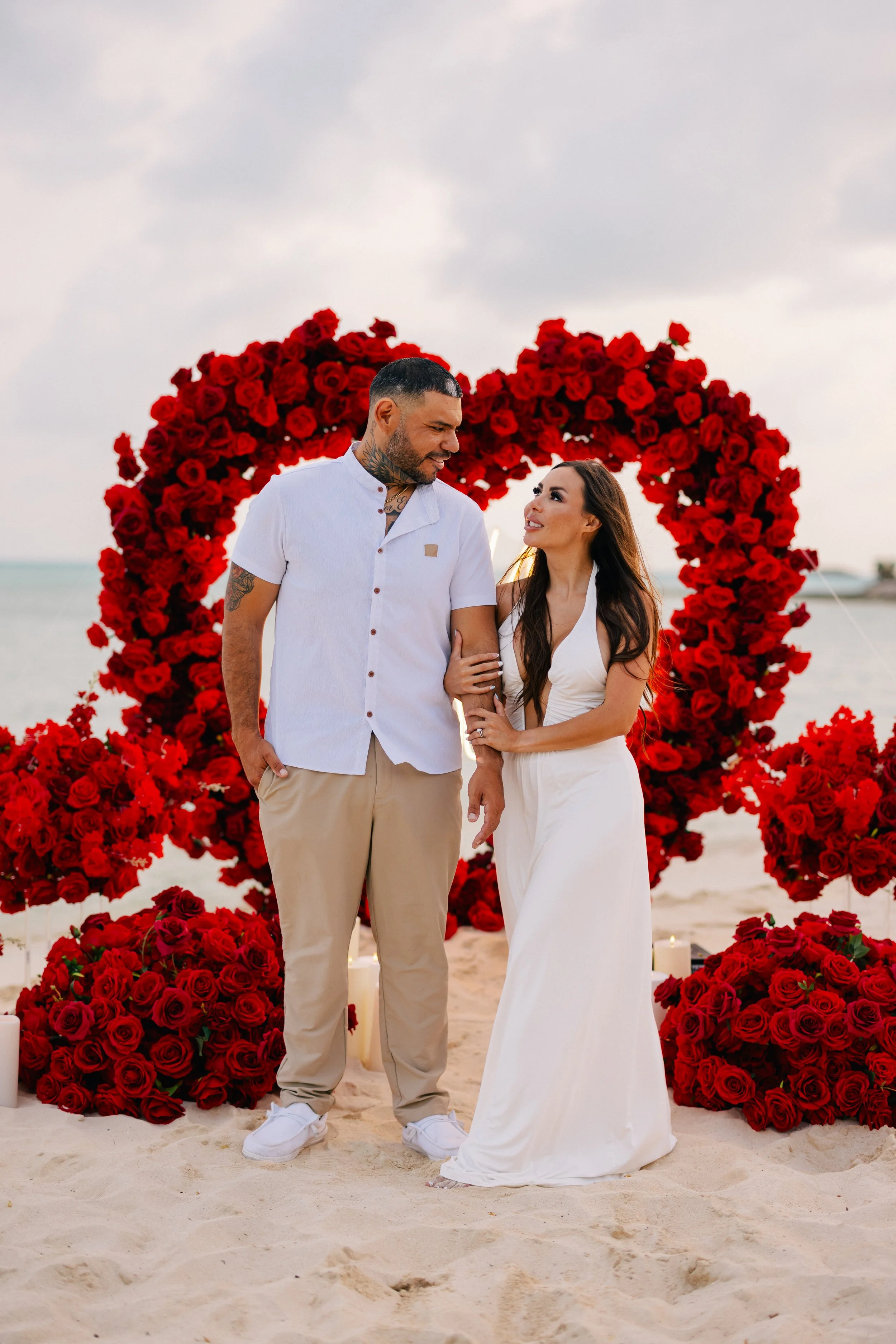 A couple standing close together on the beach, surrounded by a heart-shaped arrangement of red roses, during sunset. The man is wearing a white short-sleeved shirt and beige pants, and the woman is in a white dress. They are looking into each other's