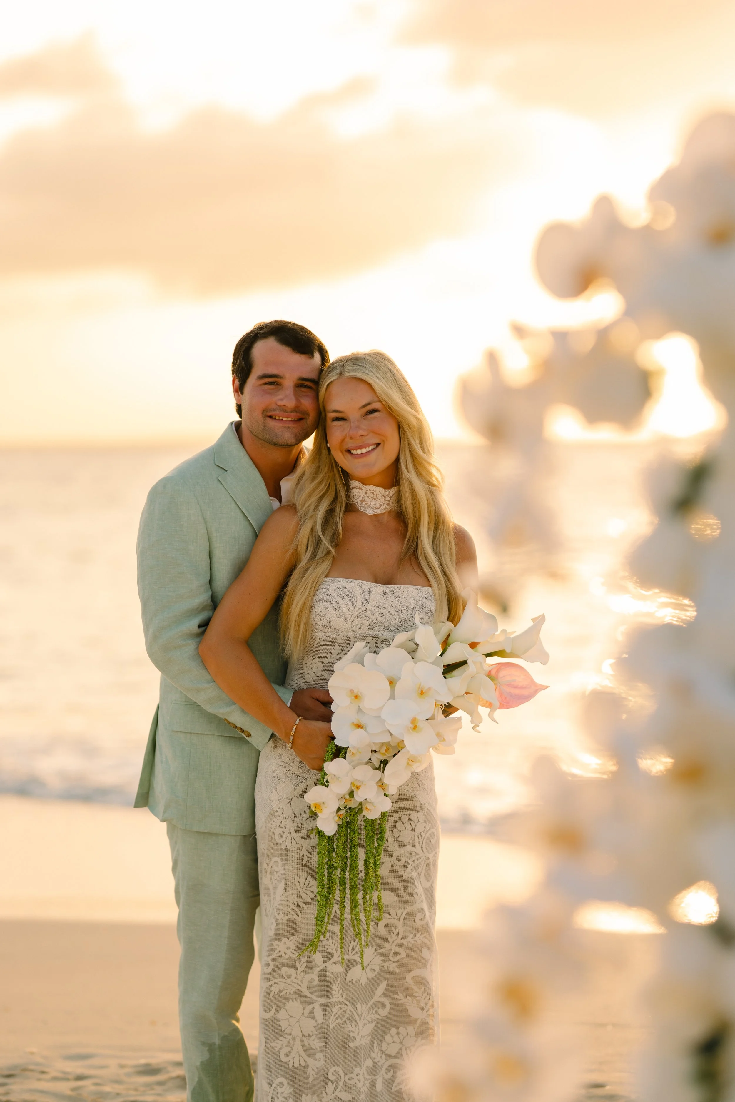 A couple in wedding attire standing on the beach at sunset, smiling with the ocean and an island in the background, holding a bouquet of white flowers.