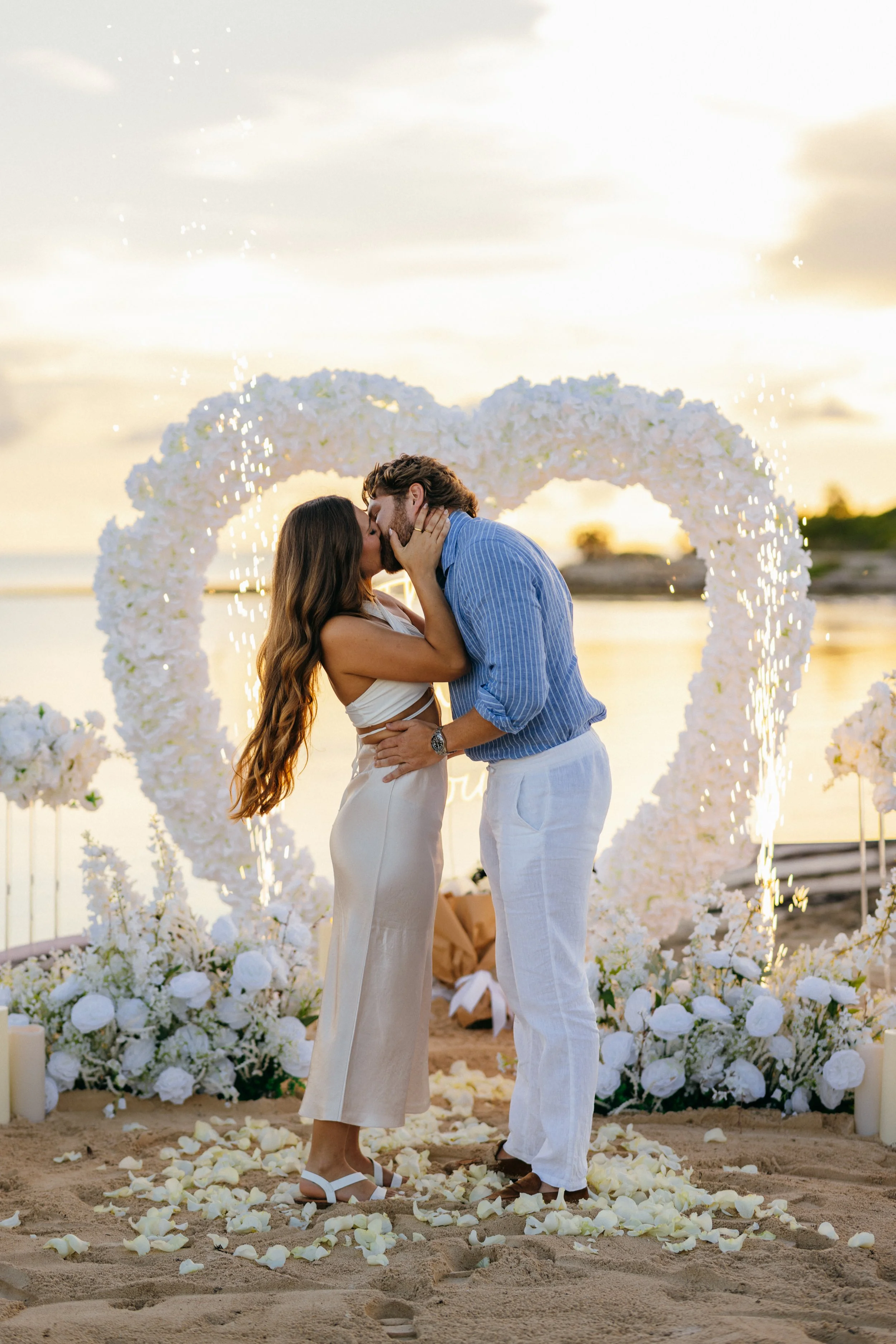 A couple sharing a kiss during a beach wedding ceremony at sunset with white flowers, rose petals on the sand, and a floral arch in the background.